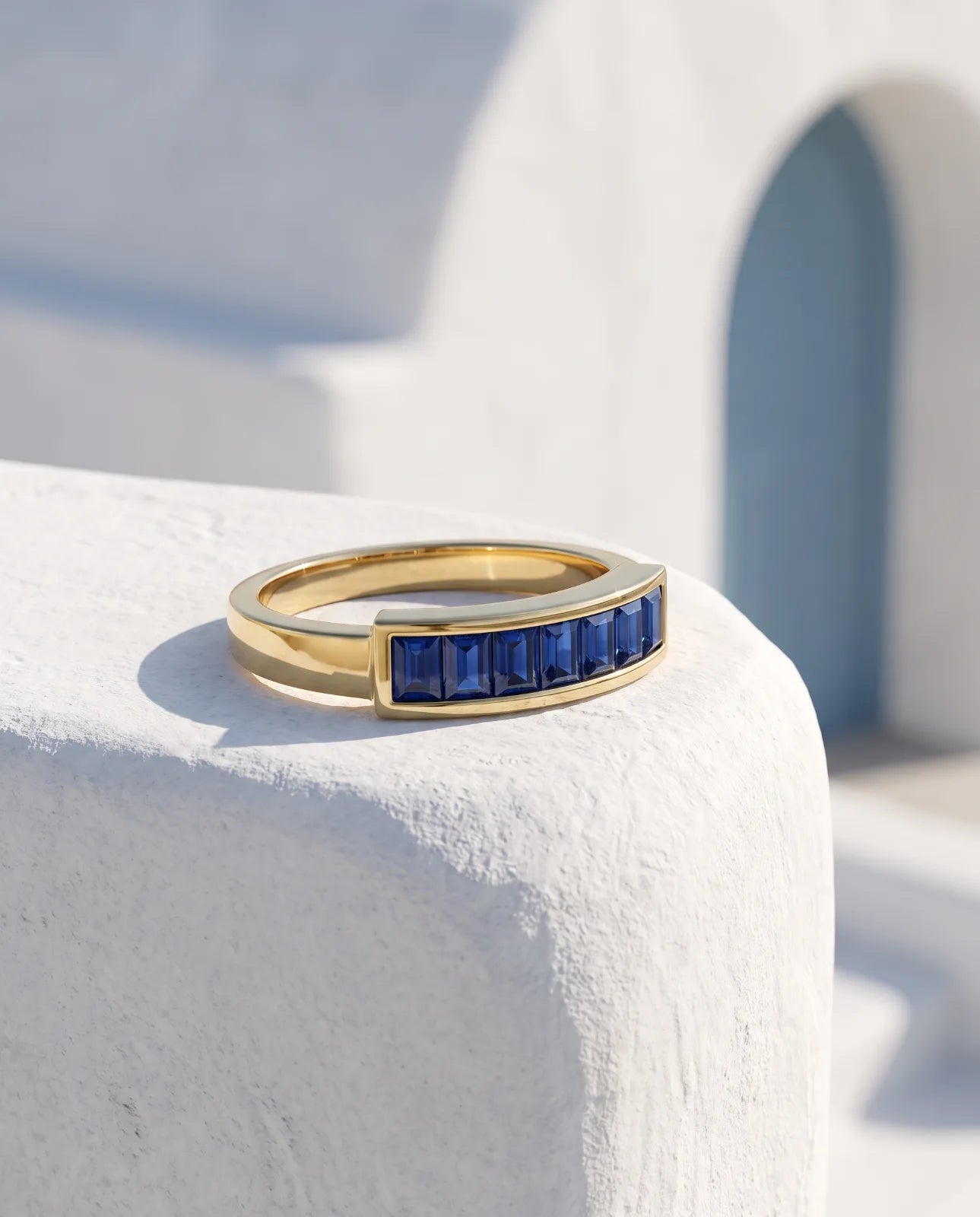 Akrotiri Band in 14k yellow gold, seven emerald-cut blue sapphires channel-set in a raised bar across a flat-top band, resting at an angle on a sunlit whitewashed Santorini ledge with a blue arched doorway behind