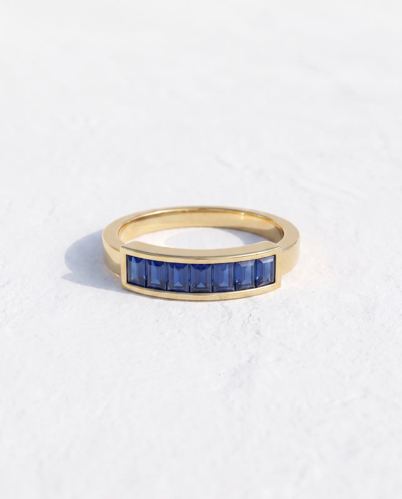 Akrotiri Band in 14k yellow gold, seven emerald-cut blue sapphires channel-set in a raised bar across a flat-top band, standing upright in a clean studio shot on a plain white background