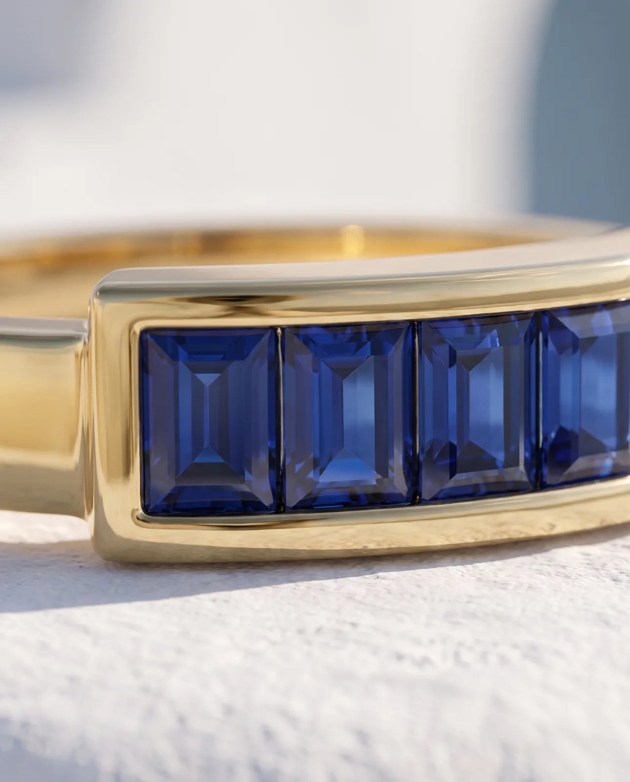 Akrotiri Band in 14k yellow gold, seven emerald-cut blue sapphires channel-set in a raised bar across a flat-top band, macro detail of the sapphires sitting flush between the heavy polished gold rails