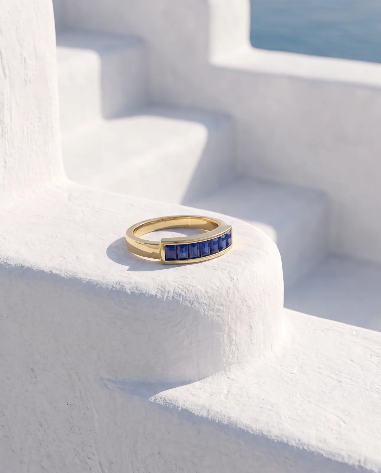 Akrotiri Band in 14k yellow gold, seven emerald-cut blue sapphires channel-set in a raised bar across a flat-top band, set on whitewashed Santorini steps with the Aegean sea beyond