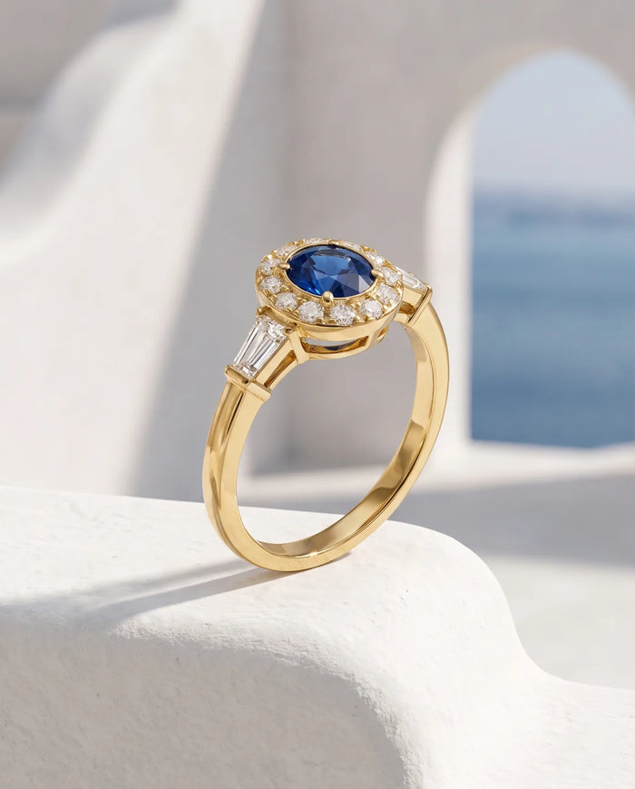 Aureole Ring in 14k yellow gold, a round blue sapphire framed by a diamond halo with a tapered baguette on each shoulder, standing upright on a sunlit whitewashed ledge with a Santorini archway and the blue Aegean behind it