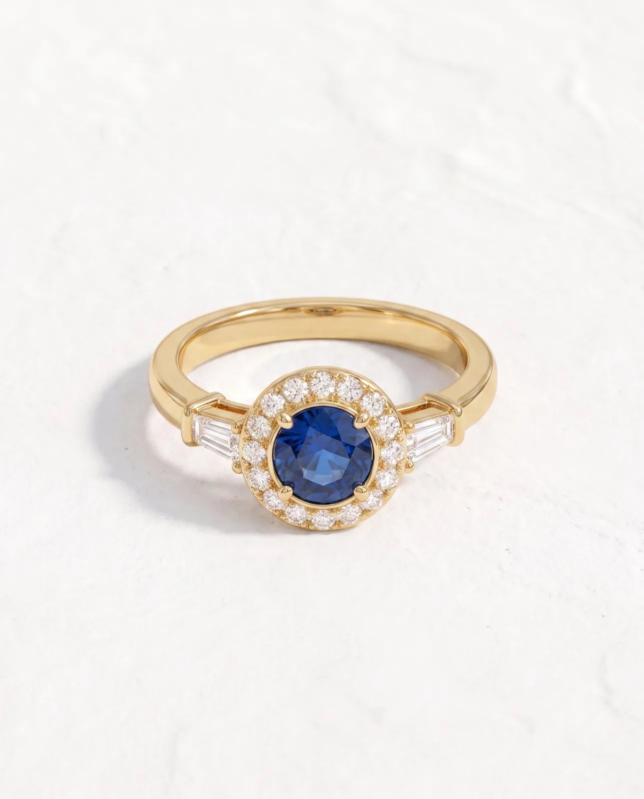 Aureole Ring in 14k yellow gold, a round blue sapphire framed by a diamond halo with a tapered baguette on each shoulder, shown from above on a plain white studio background