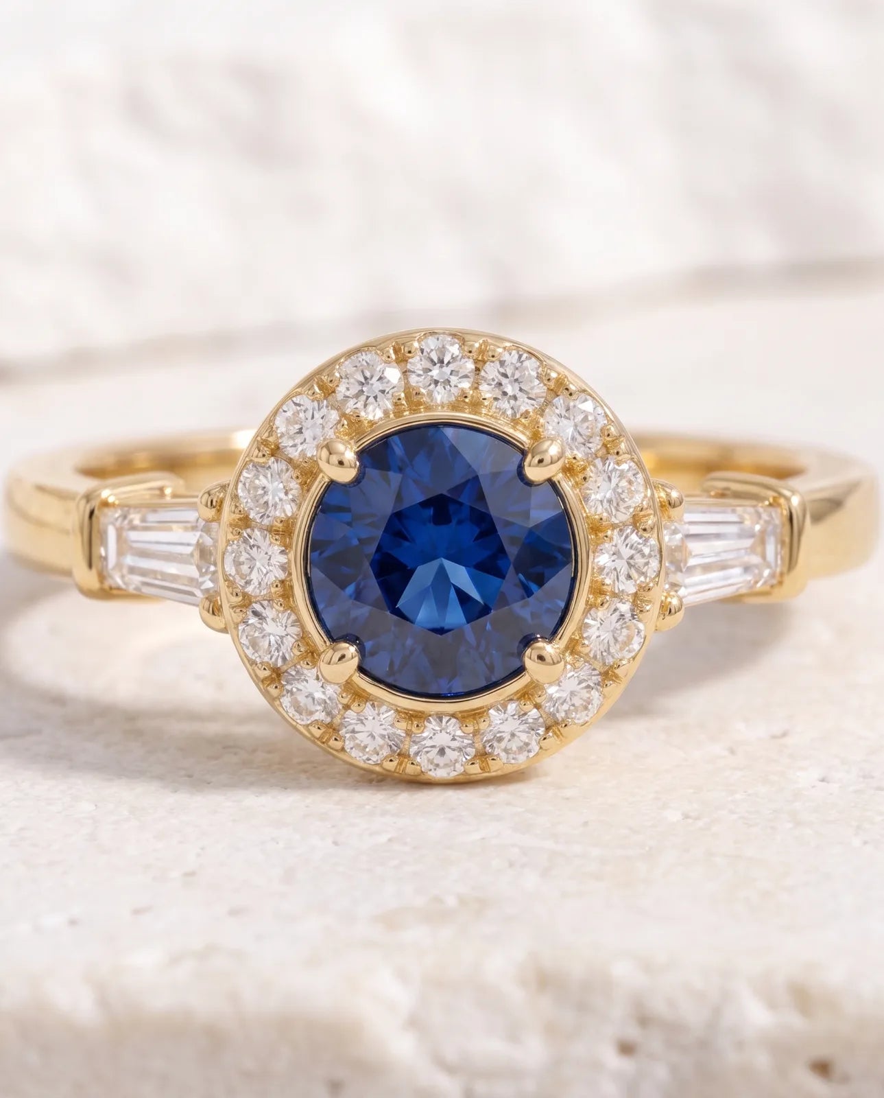 Aureole Ring in 14k yellow gold, a round blue sapphire framed by a diamond halo with a tapered baguette on each shoulder, in a macro close-up showing the four prongs, the full circle of pave diamonds and the bar-set baguettes