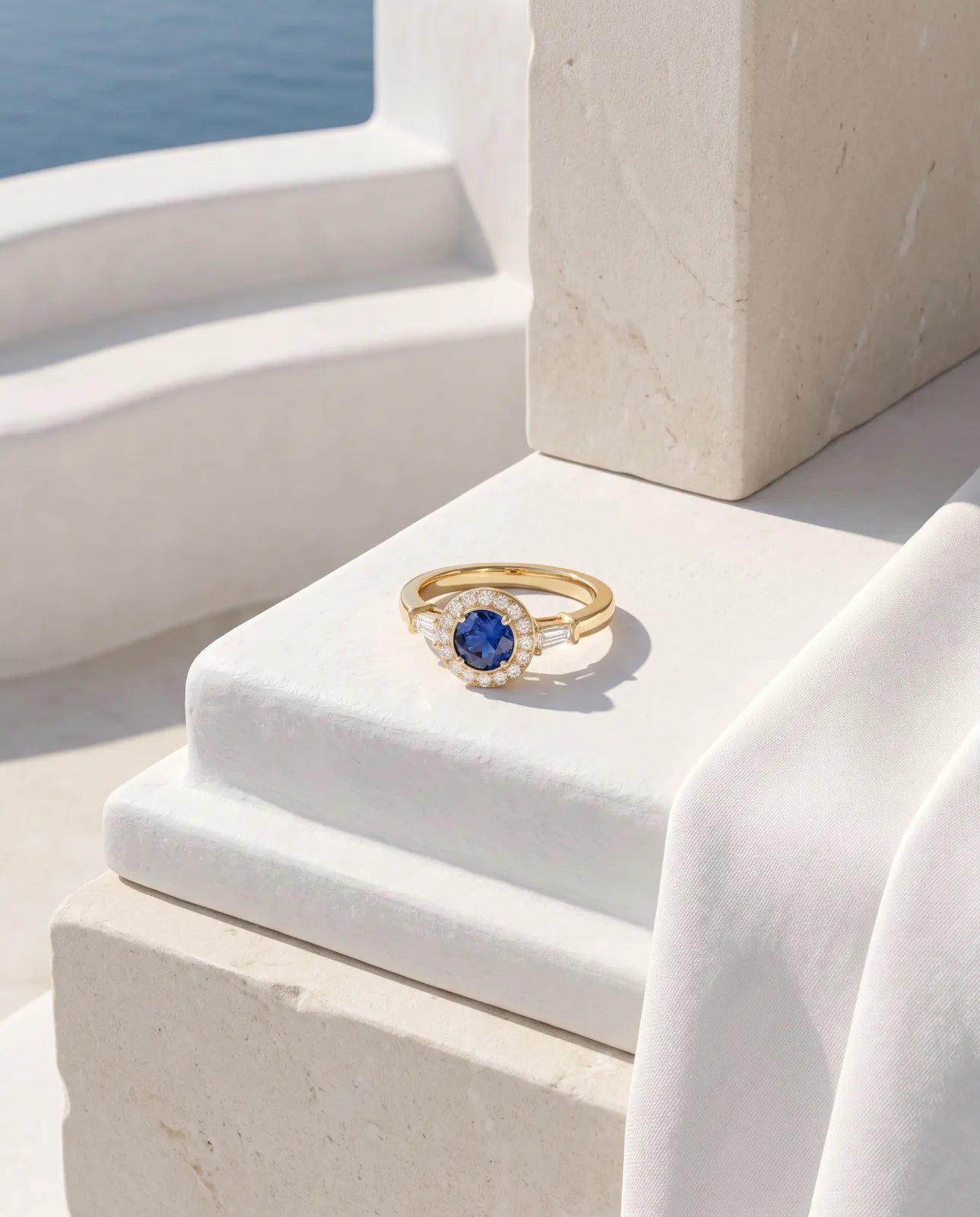Aureole Ring in 14k yellow gold, a round blue sapphire framed by a diamond halo with a tapered baguette on each shoulder, resting on a white plaster step beside draped linen with the sea in the distance