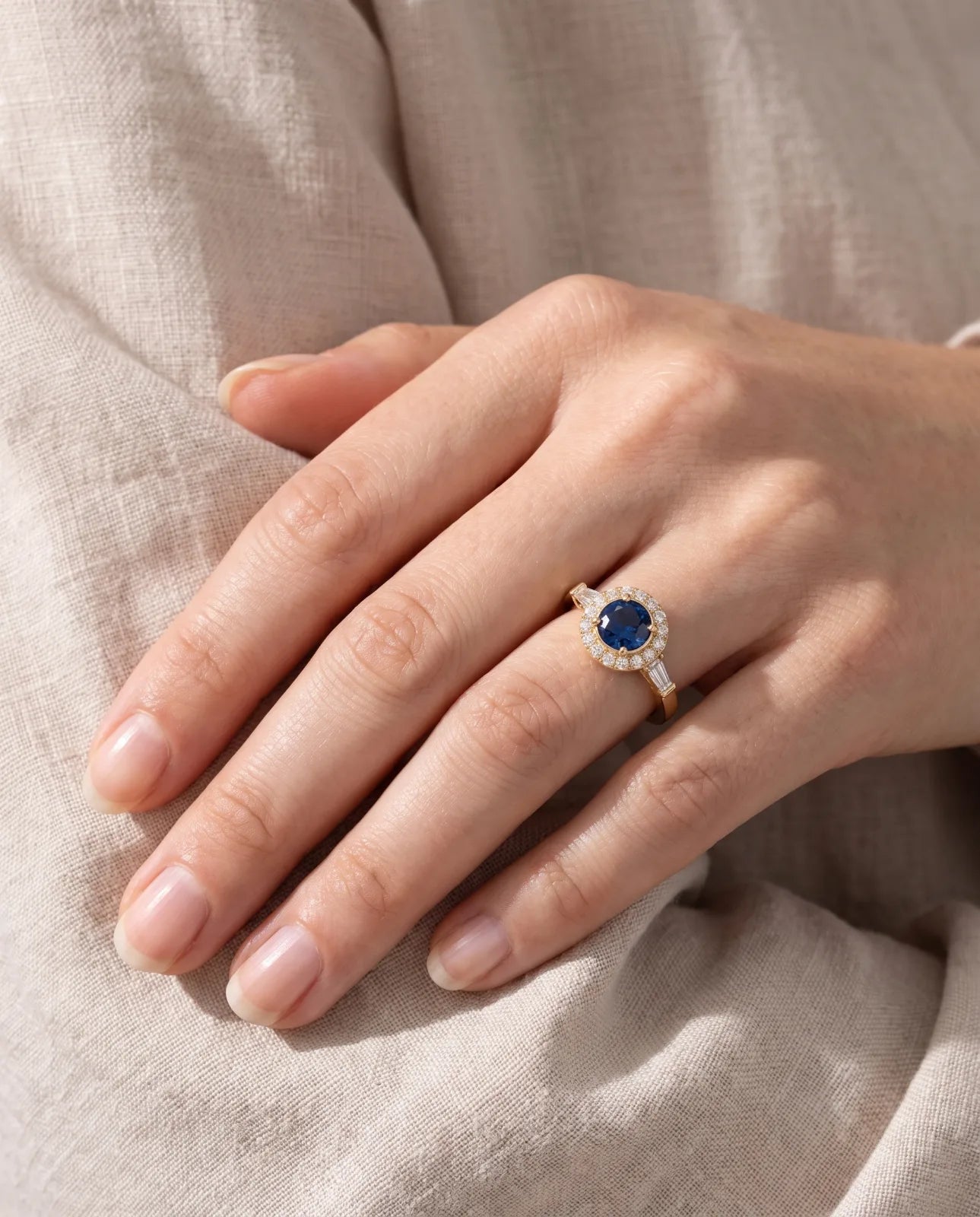 Aureole Ring in 14k yellow gold, a round blue sapphire framed by a diamond halo with a tapered baguette on each shoulder, worn on a hand resting on soft oatmeal linen in warm natural light