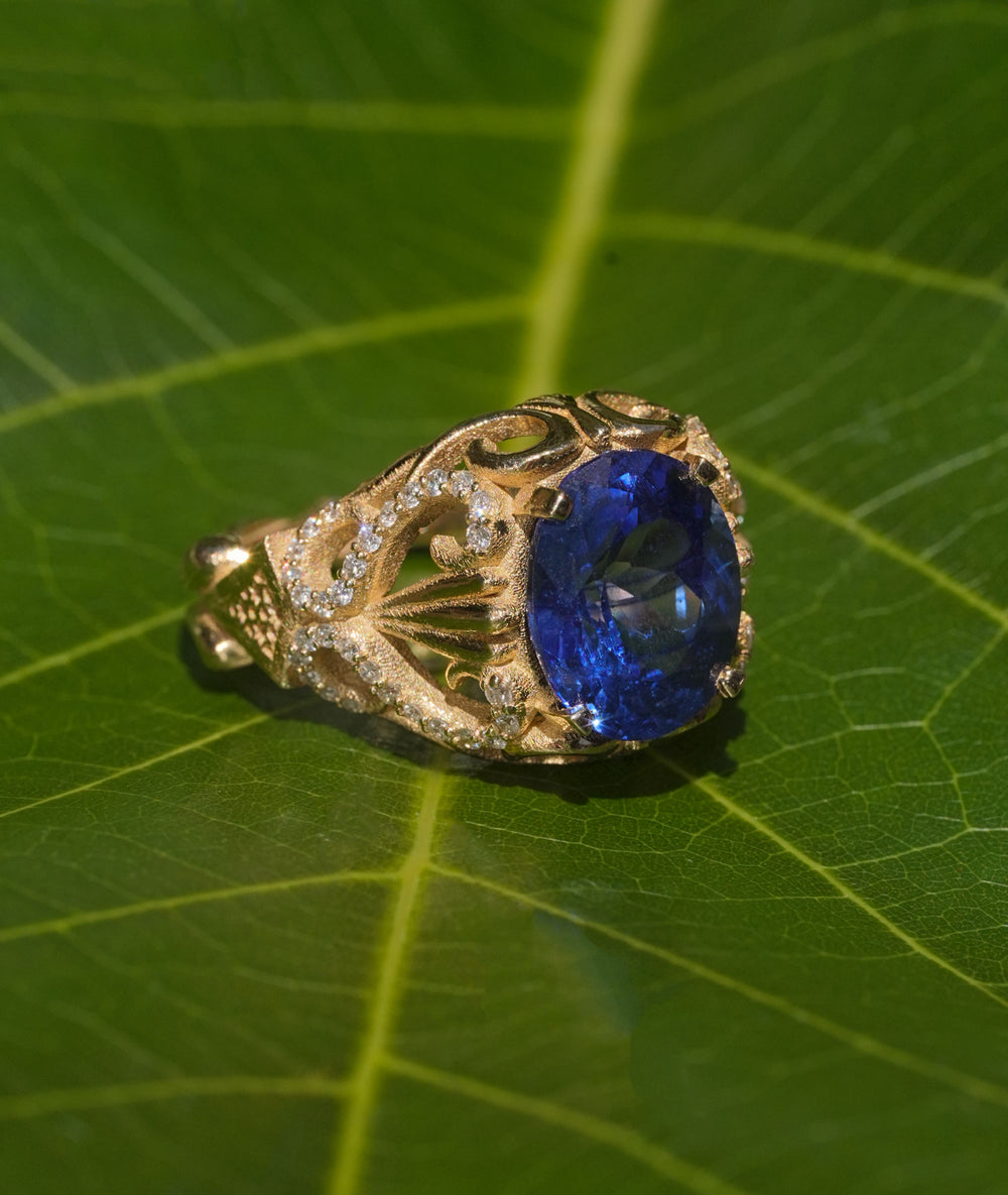 Gold ring with an oval blue sapphire center stone, intricate filigree design, and diamond accents, inspired by Mediterranean elegance.