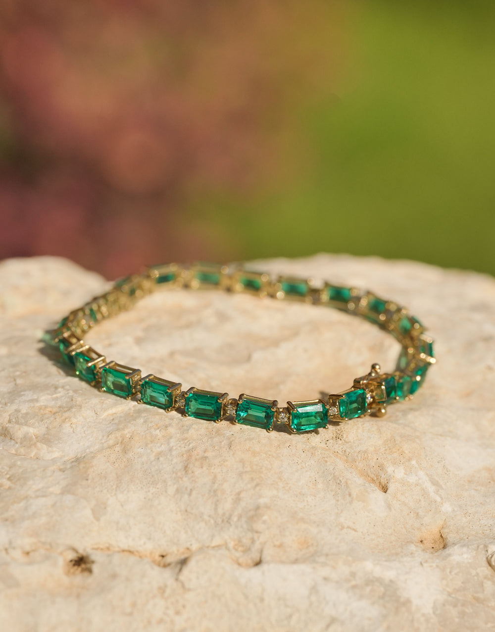Gold tennis bracelet featuring rectangular emerald gemstones and diamond accents, worn on a wrist against a soft turquoise background.