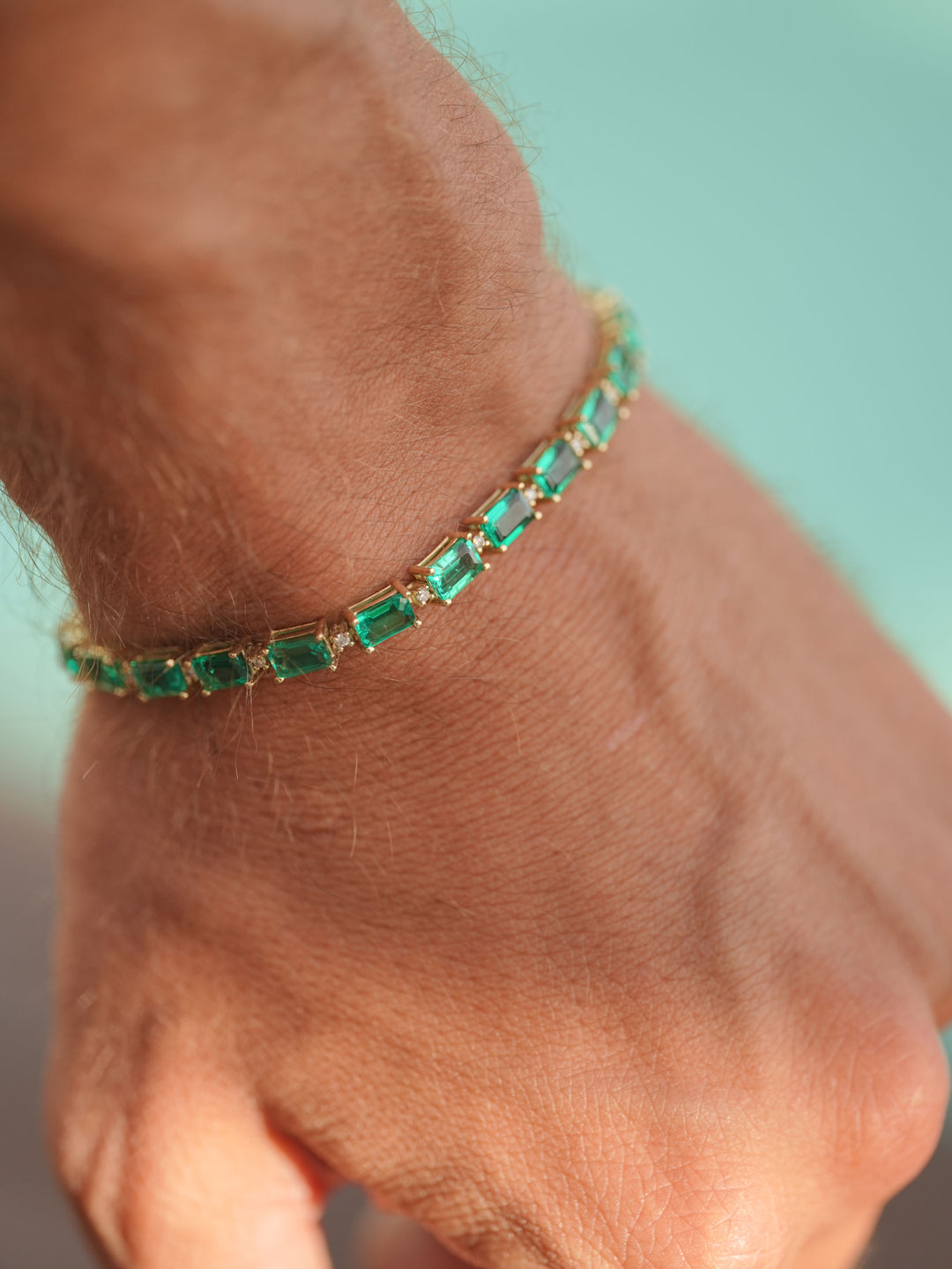Gold tennis bracelet featuring rectangular emerald gemstones and diamond accents, worn on a wrist against a soft turquoise background.