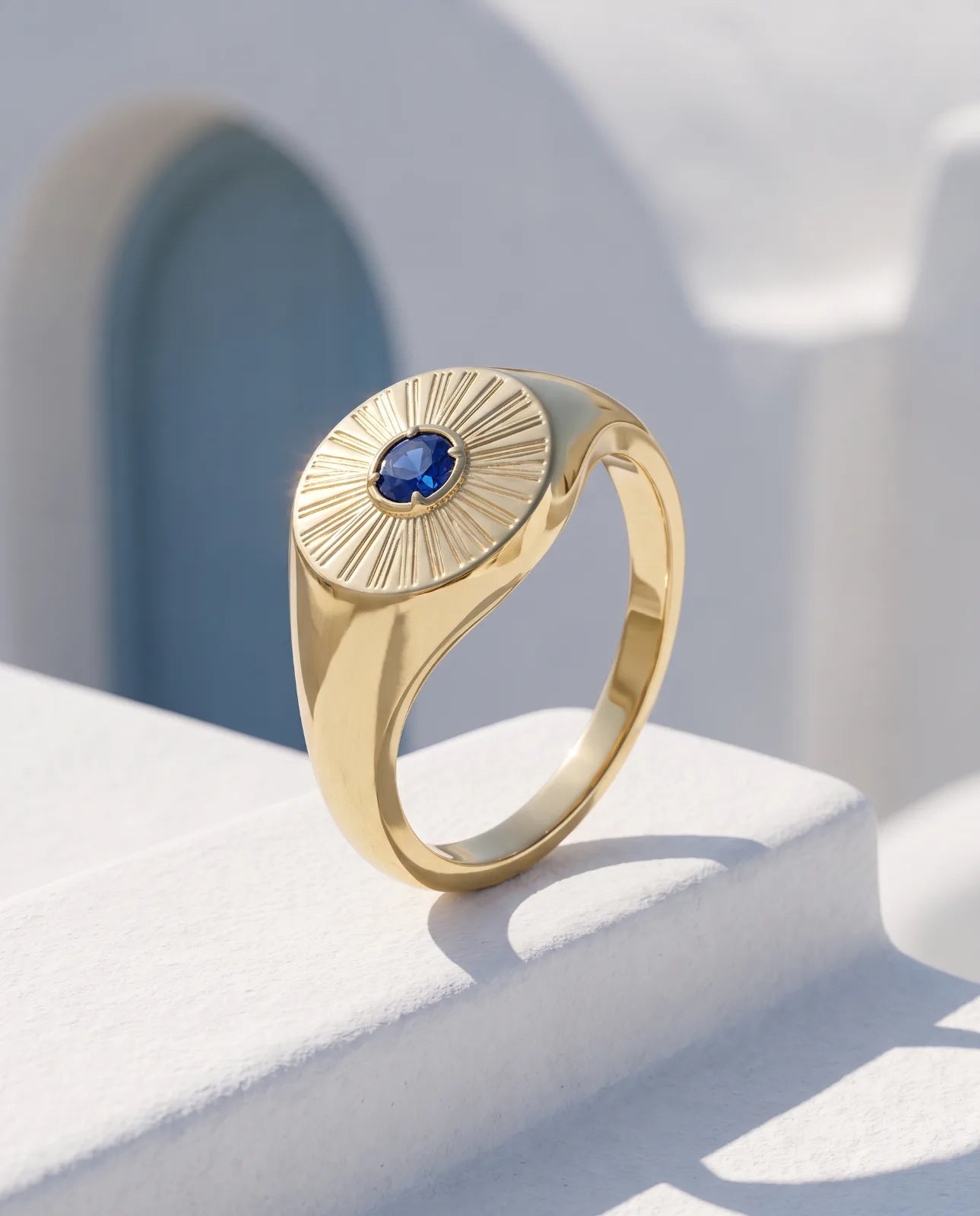 Helia Signet in 14k yellow gold with a blue sapphire at the centre of an engraved sunburst face, standing upright on a sunlit whitewashed ledge against a blue arched Santorini doorway.