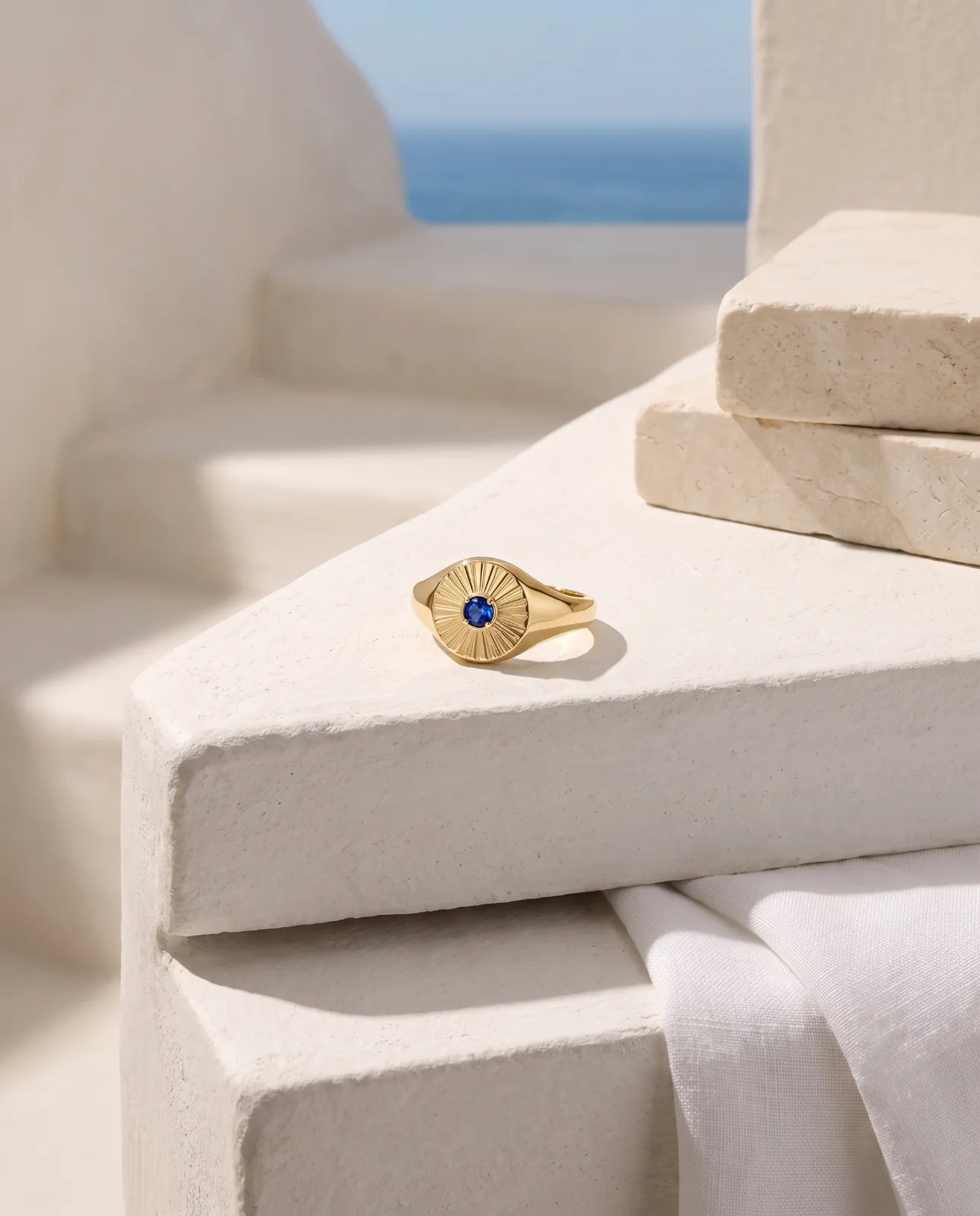 Helia Signet in 14k yellow gold with a blue sapphire at the centre of an engraved sunburst face, lying face-up on a whitewashed ledge beside stacked limestone tiles and white linen, with the Aegean sea behind.