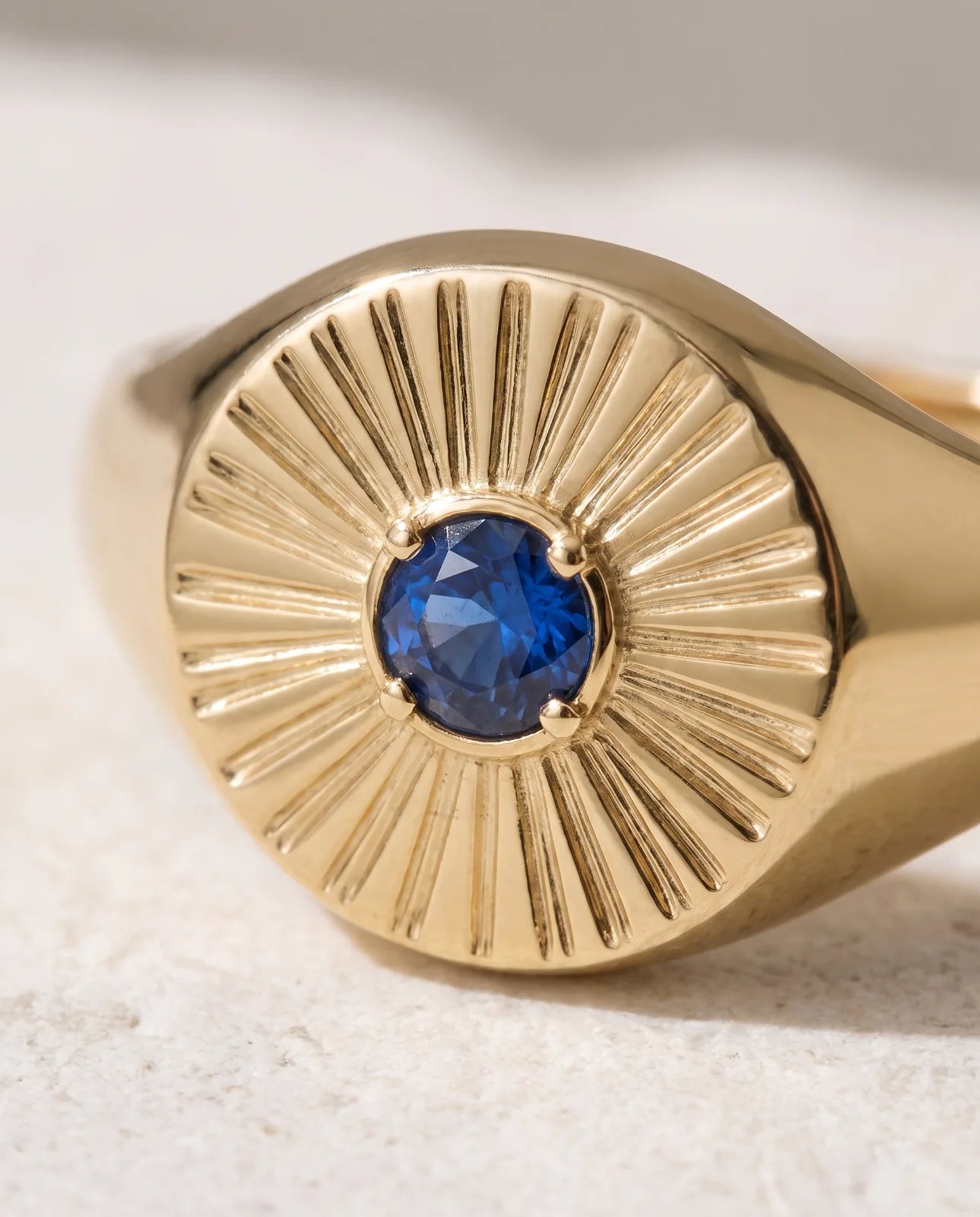 Helia Signet in 14k yellow gold with a blue sapphire at the centre of an engraved sunburst face, shown in macro close-up so the four-prong setting and fine radiating ray engraving fill the frame.