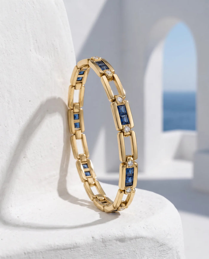Kastro Bracelet in 14k yellow gold, alternating open rectangular links with channel-set blue sapphire bars capped by bezel diamonds, standing upright on a whitewashed plinth against a sunlit Santorini archway framed on the Aegean