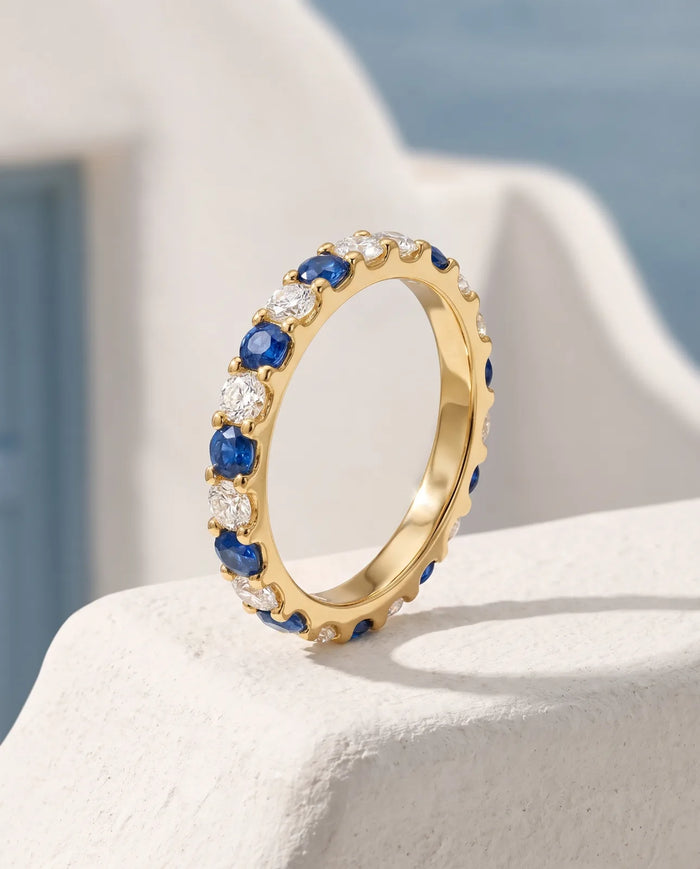 Kyma Eternity Band in 14k yellow gold, a full eternity band alternating round blue sapphires and white diamonds, standing upright on a sunlit whitewashed ledge with a blue shutter and the Aegean sea blurred behind