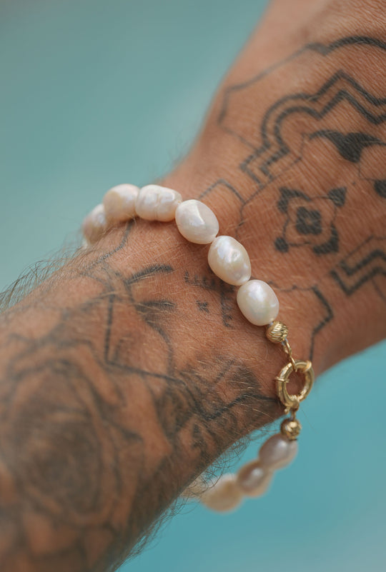 Pearl bracelet with irregular freshwater pearls and a gold clasp, worn on a tattooed wrist against a turquoise background.