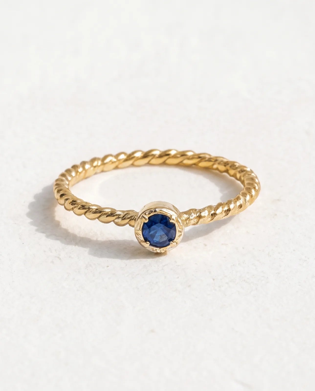 Meltemi Ring in 14k yellow gold, a round blue sapphire on a twisted rope band, laid flat on a soft off-white studio background showing the full rope-twist shank and bezel-with-prongs cup