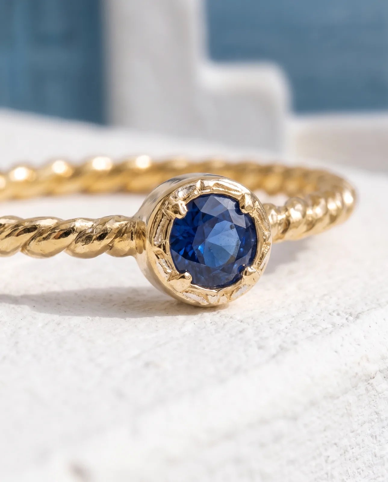 Meltemi Ring in 14k yellow gold, a round blue sapphire on a twisted rope band, macro detail of the faceted stone in its bezel-with-prongs setting against a whitewashed ledge and deep blue Aegean water