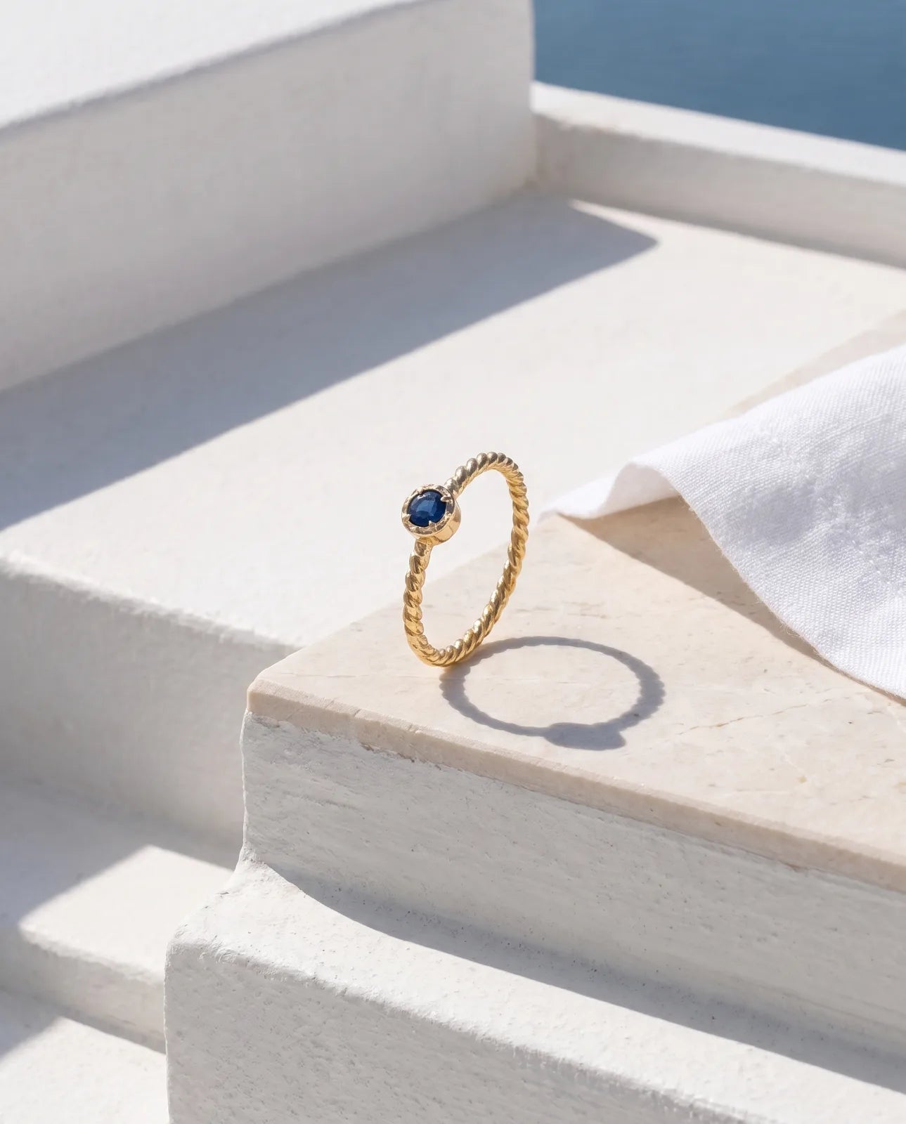 Meltemi Ring in 14k yellow gold, a round blue sapphire on a twisted rope band, standing on a sunlit limestone step beside white linen with the sea beyond, casting a crisp circular shadow
