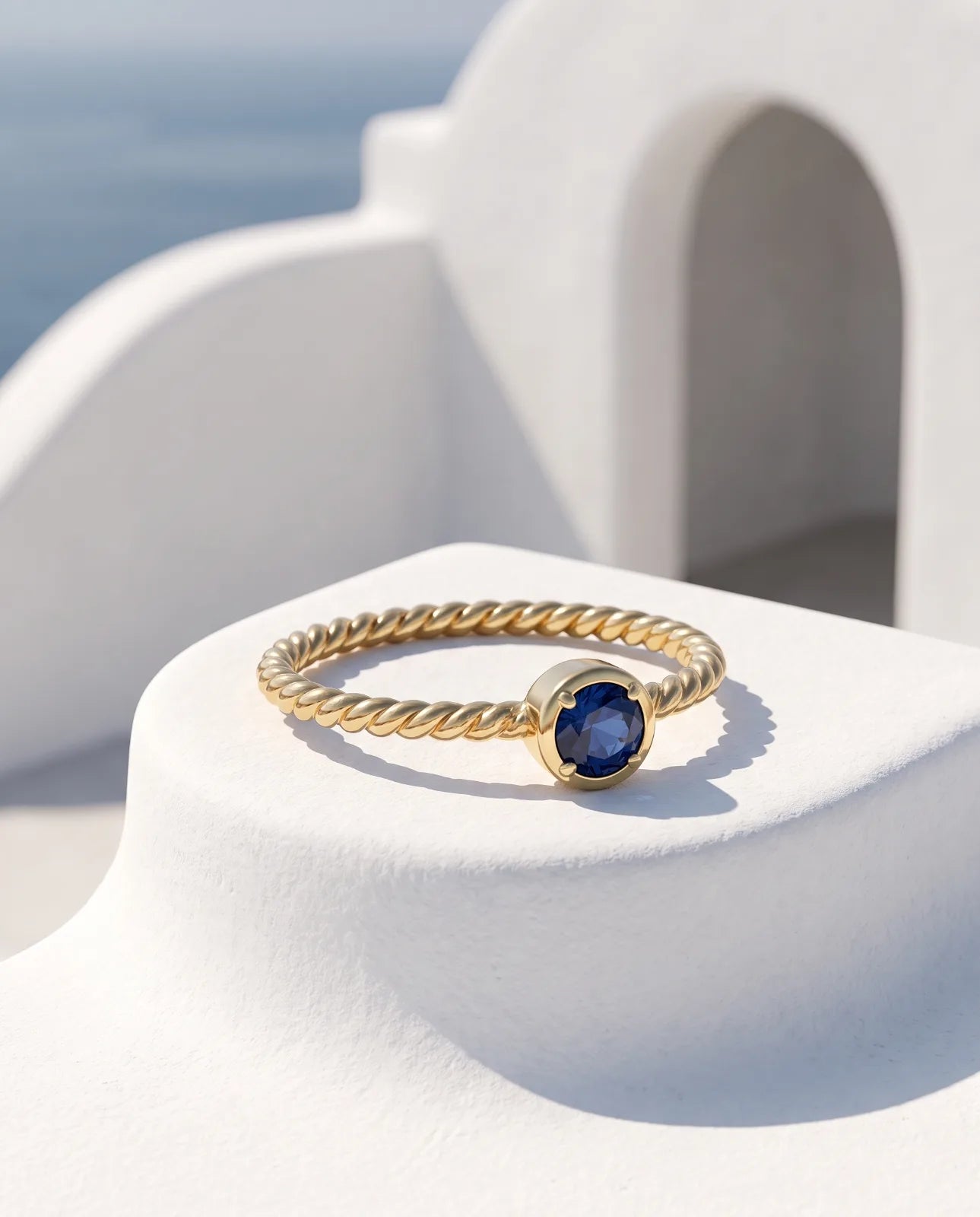 Meltemi Ring in 14k yellow gold, a round blue sapphire on a twisted rope band, resting at an angle on a curved white plaster pedestal with a Cycladic arch and the sea softly out of focus behind