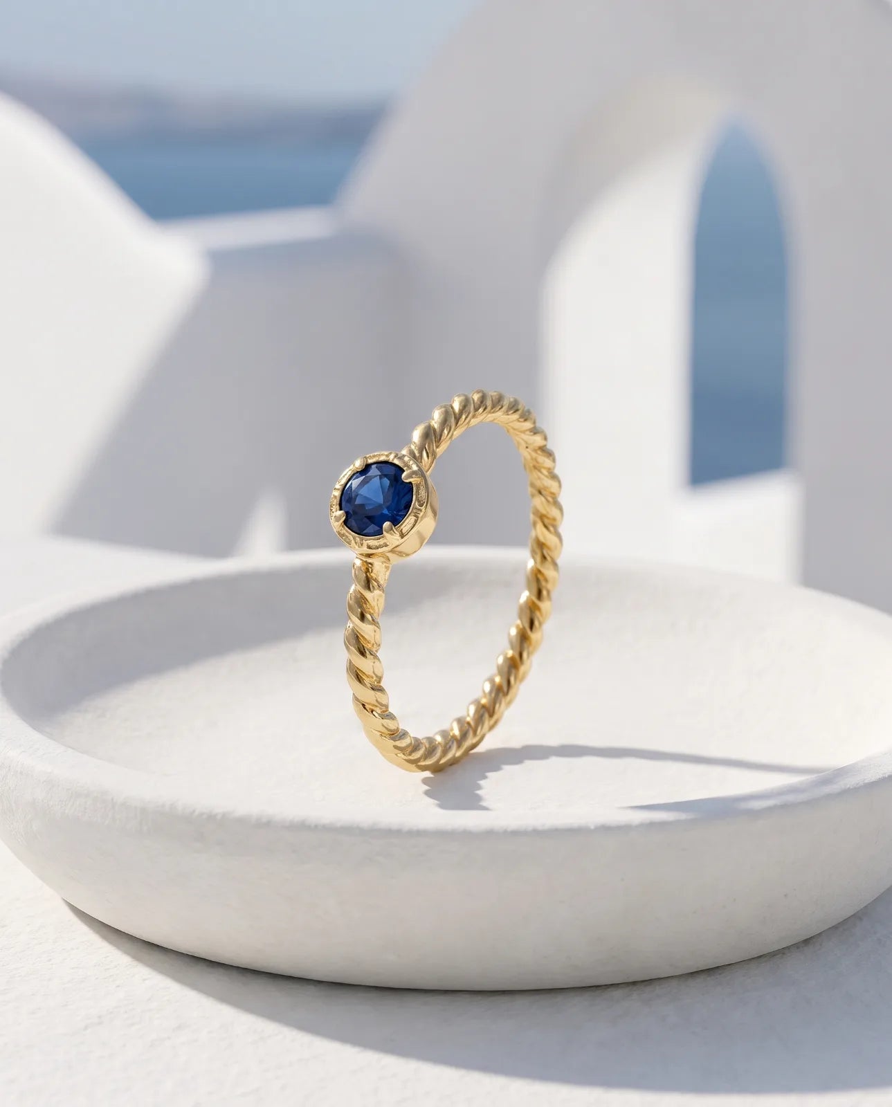 Meltemi Ring in 14k yellow gold, a round blue sapphire on a twisted rope band, standing upright in a white ceramic dish with a sunlit Santorini whitewashed archway and the Aegean sea blurred behind