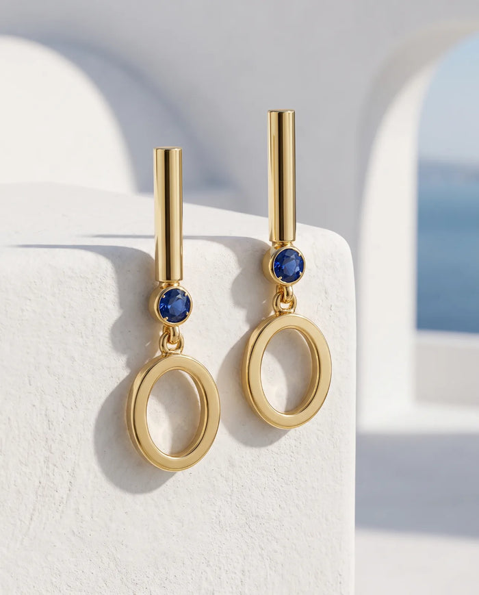 Oia Drops in 14k yellow gold, bar-and-circle drop earrings with a bezel-set blue sapphire above a swinging open O, the pair angled against a sunlit whitewashed ledge with a blue Aegean arch behind.