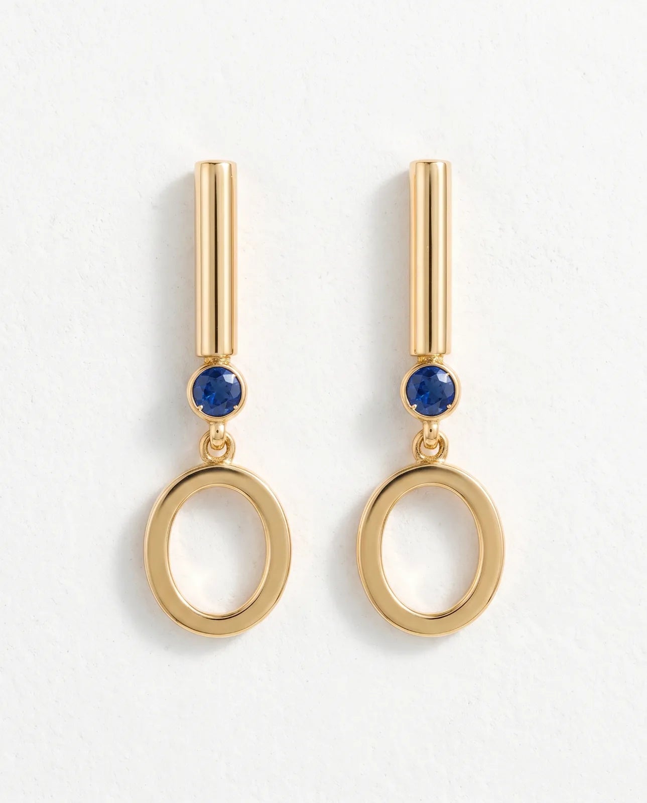 Oia Drops in 14k yellow gold, bar-and-circle drop earrings with a bezel-set blue sapphire above a swinging open O, shown straight on as a matched pair on a plain white background.