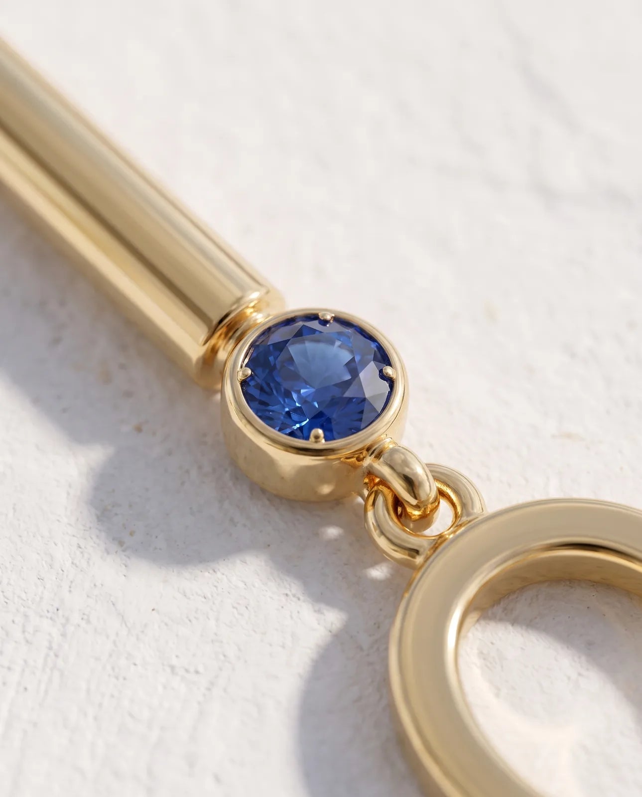 Oia Drops in 14k yellow gold, bar-and-circle drop earrings with a bezel-set blue sapphire above a swinging open O, in macro close-up on the bezel setting, jump ring and polished bar.