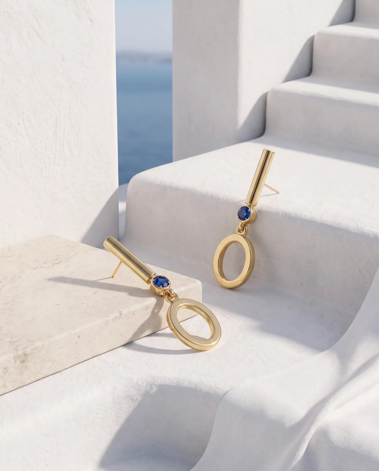 Oia Drops in 14k yellow gold, bar-and-circle drop earrings with a bezel-set blue sapphire above a swinging open O, laid across whitewashed Santorini steps and a limestone block with the sea beyond.