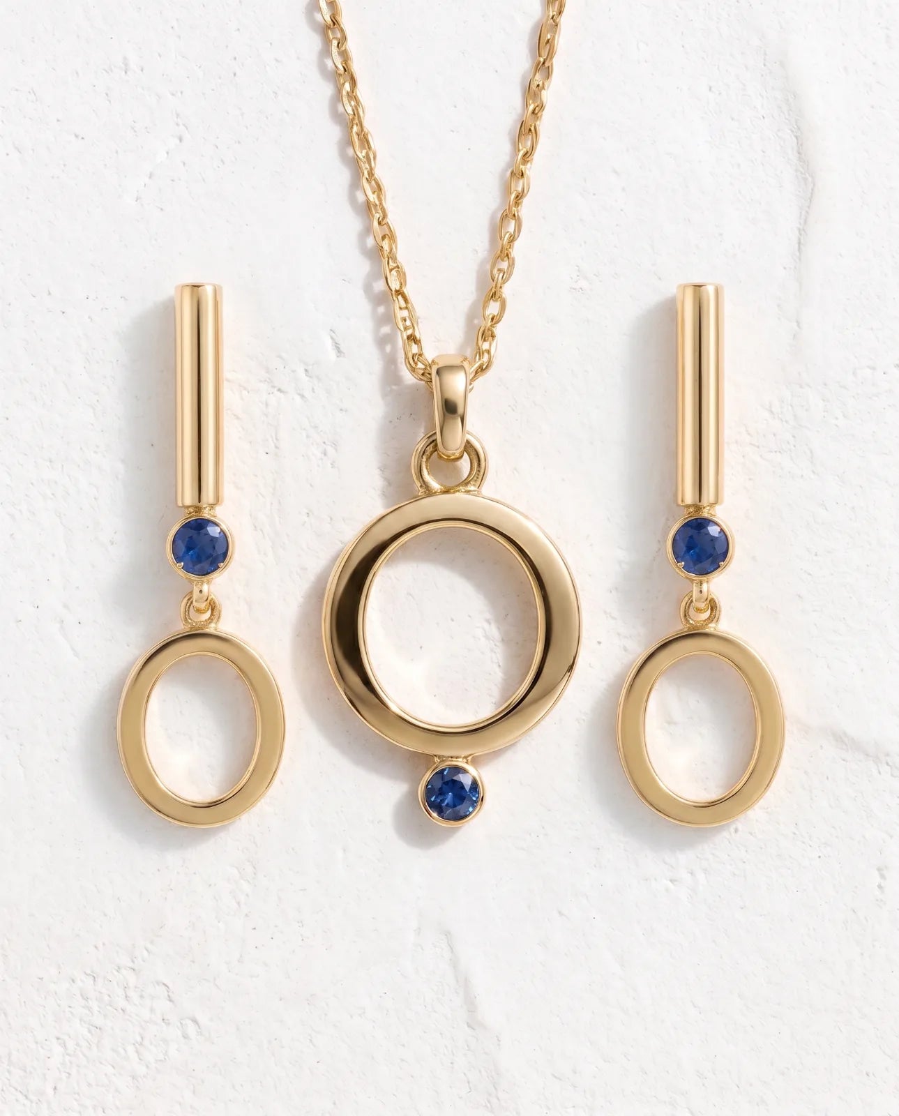 Oia Drops in 14k yellow gold, bar-and-circle drop earrings with a bezel-set blue sapphire above a swinging open O, shown as a set flanking the matching Oia Pendant on its cable chain.