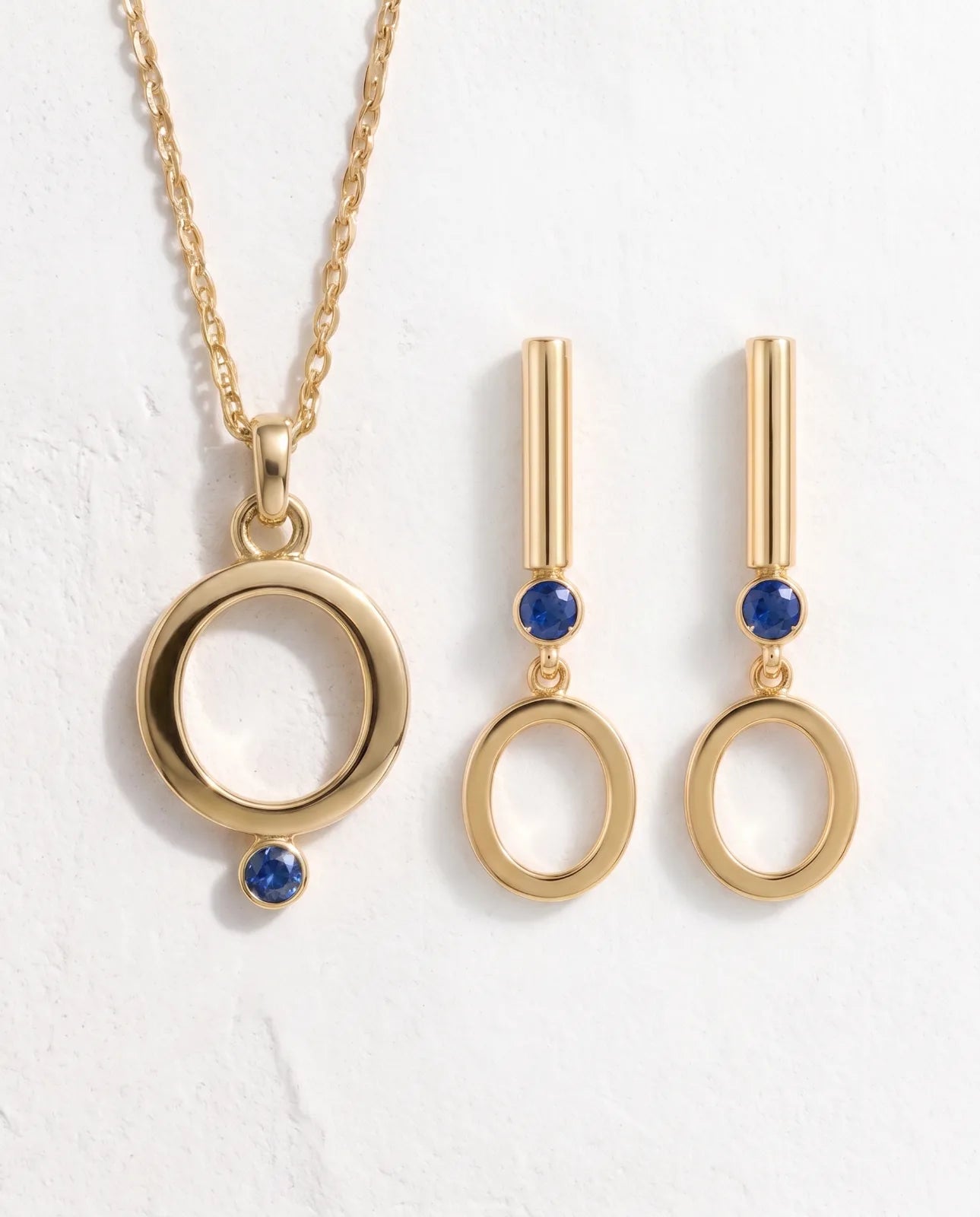 Oia Drops in 14k yellow gold, bar-and-circle drop earrings with a bezel-set blue sapphire above a swinging open O, arranged as a set beside the matching Oia Pendant on a white plaster surface.