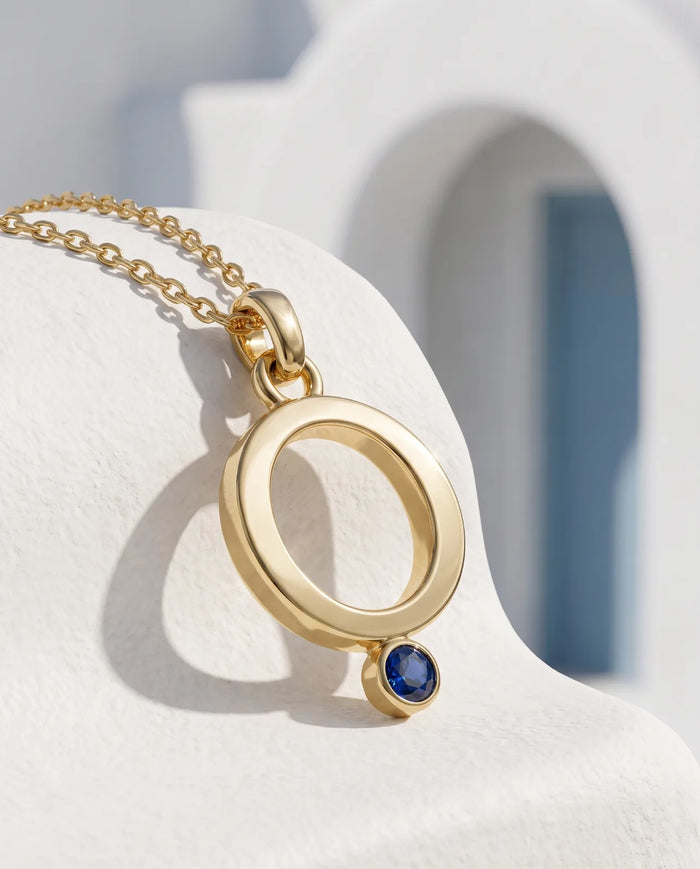 Oia Pendant in 14k yellow gold, an open circle with a bezel-set blue sapphire at its base on a fine cable chain, shown angled against a sunlit whitewashed Santorini wall with a blue doorway behind.