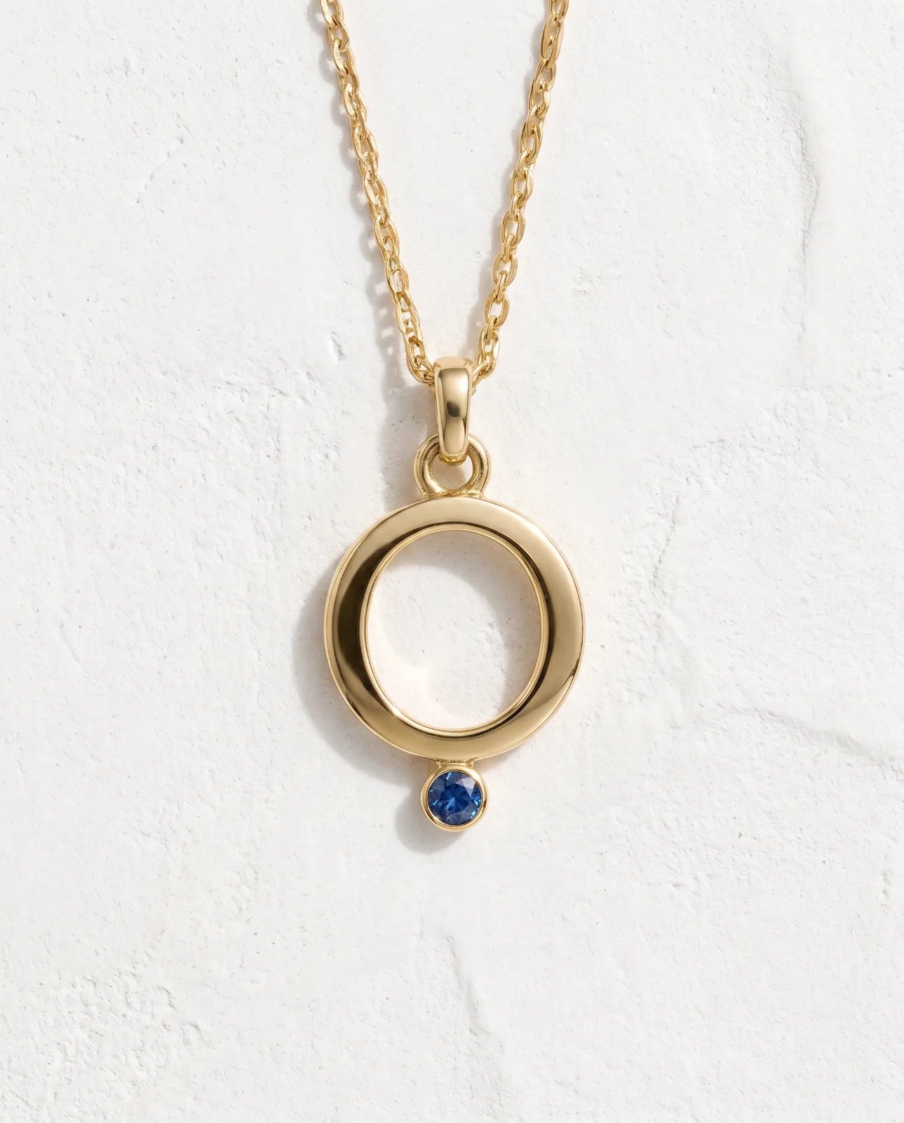 Oia Pendant in 14k yellow gold, an open circle with a bezel-set blue sapphire at its base on a fine cable chain, laid flat face-on against white plaster to show the full necklace and bail.