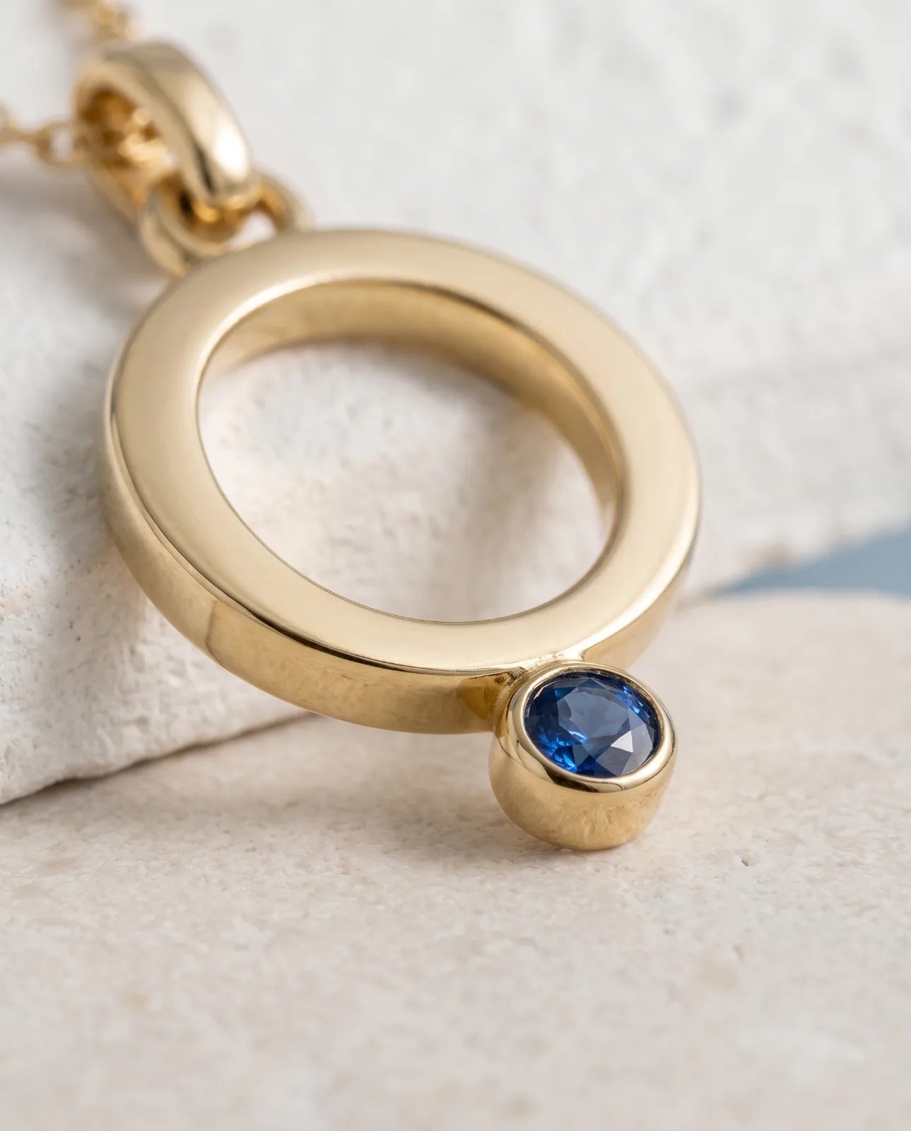 Oia Pendant in 14k yellow gold, an open circle with a bezel-set blue sapphire at its base on a fine cable chain, in a close macro view of the rounded gold profile and faceted sapphire bezel.