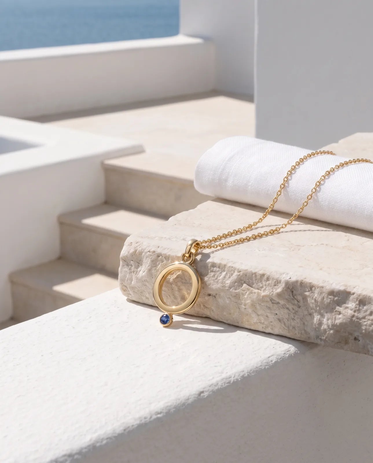 Oia Pendant in 14k yellow gold, an open circle with a bezel-set blue sapphire at its base on a fine cable chain, styled on a limestone ledge above white terrace steps with the Aegean sea beyond.