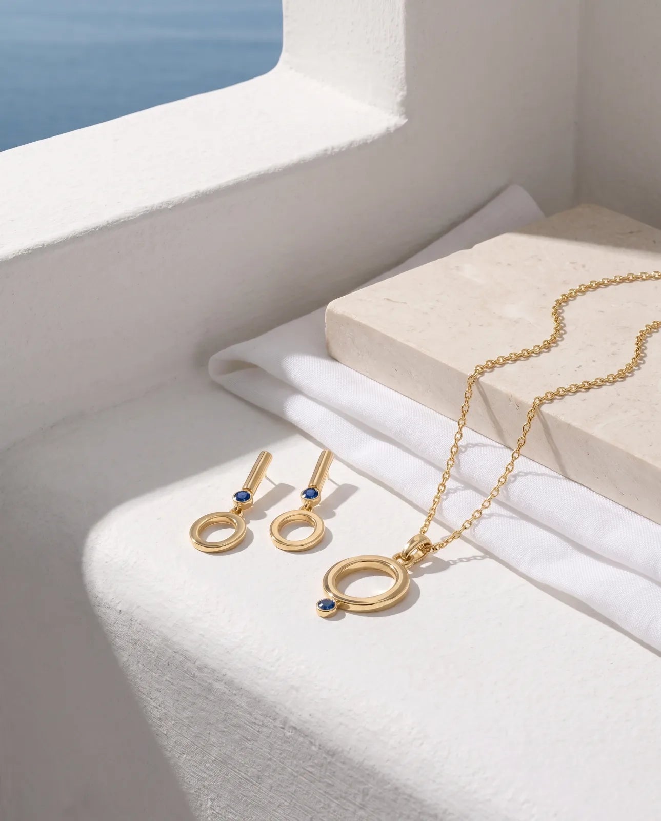 Oia Pendant in 14k yellow gold, an open circle with a bezel-set blue sapphire at its base on a fine cable chain, styled as a set with the matching Oia Drops earrings on a sunlit white ledge overlooking the sea.