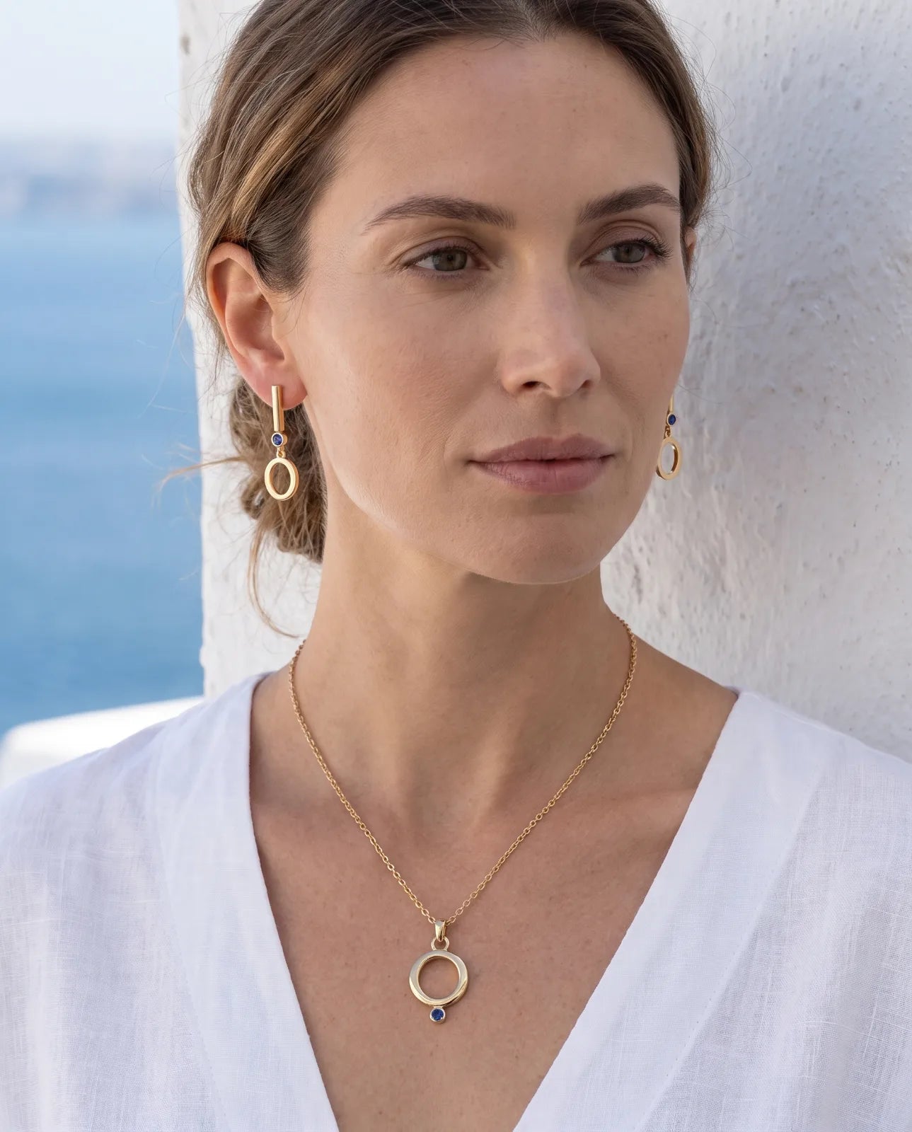 Oia Pendant in 14k yellow gold, an open circle with a bezel-set blue sapphire at its base on a fine cable chain, worn on-model as a set with the matching Oia Drops earrings against a whitewashed wall by the sea.