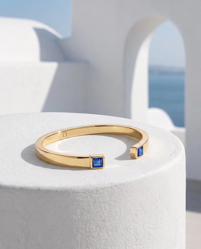 Portico Cuff in 14k yellow gold, a flat polished open cuff with a square step-cut blue sapphire set into each terminal, resting on a whitewashed plinth with a Santorini archway and the Aegean sea behind