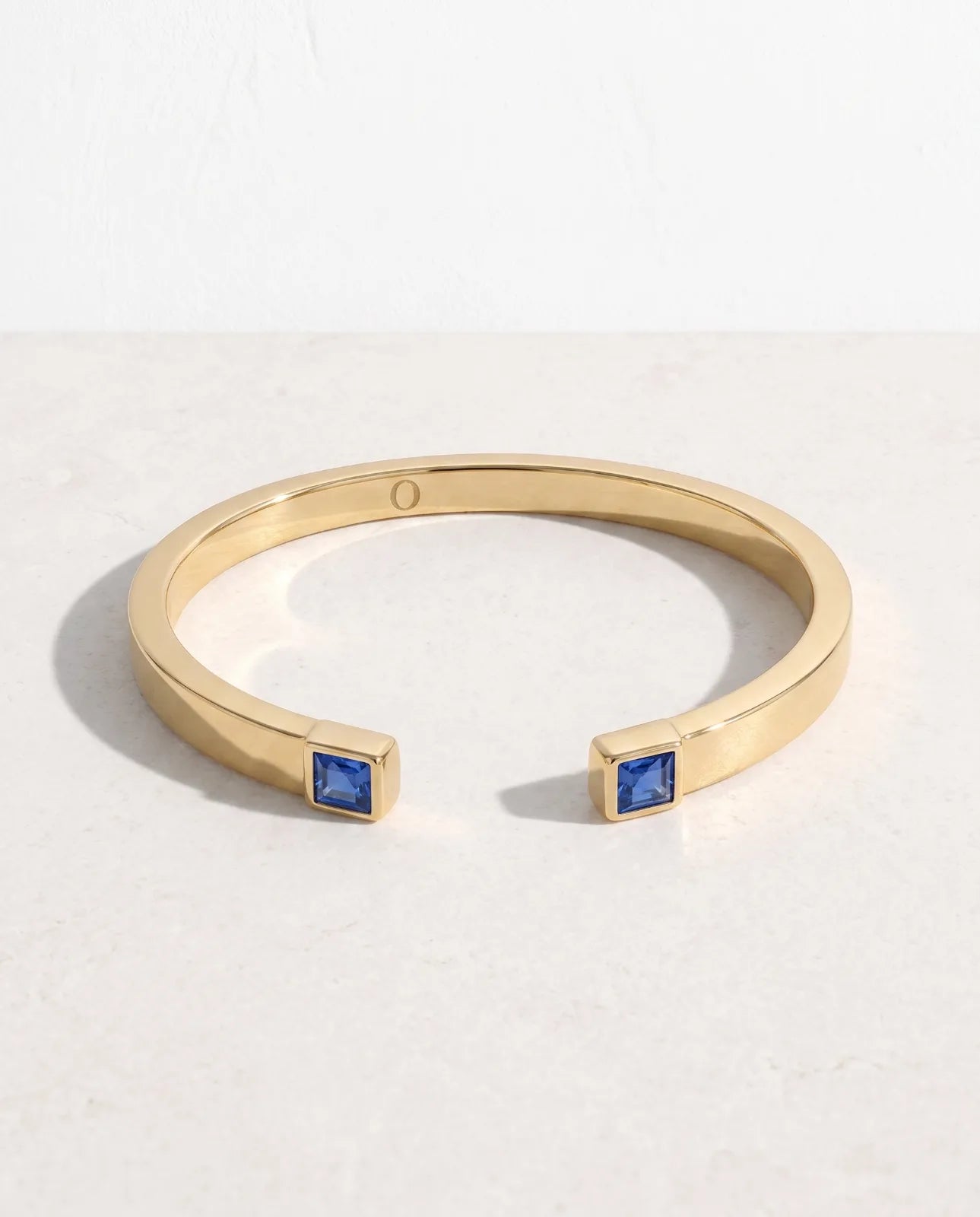 Portico Cuff in 14k yellow gold, a flat polished open cuff with a square step-cut blue sapphire set into each terminal, shown front-on against a plain white studio background with the O engraved inside the band