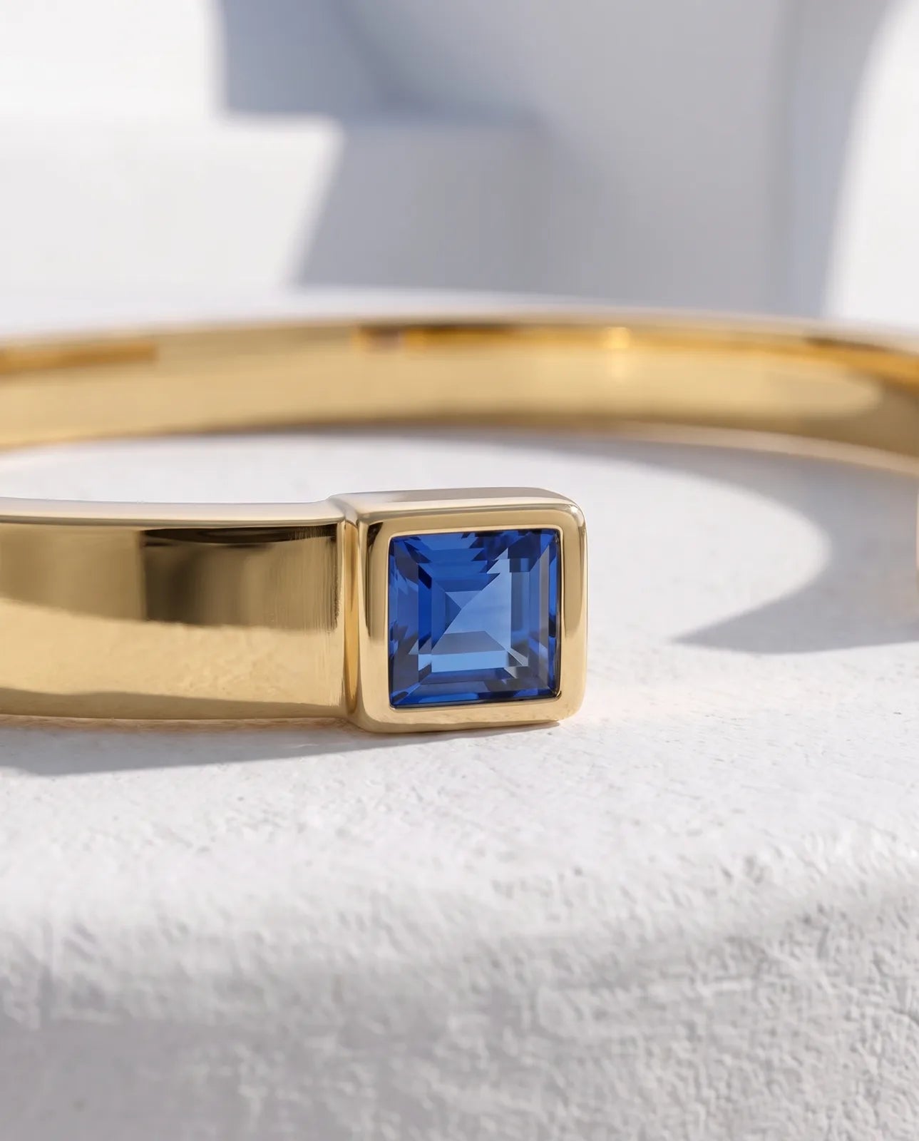 Portico Cuff in 14k yellow gold, a flat polished open cuff with a square step-cut blue sapphire set into each terminal, in macro detail showing one bezel-set sapphire flush against the polished band