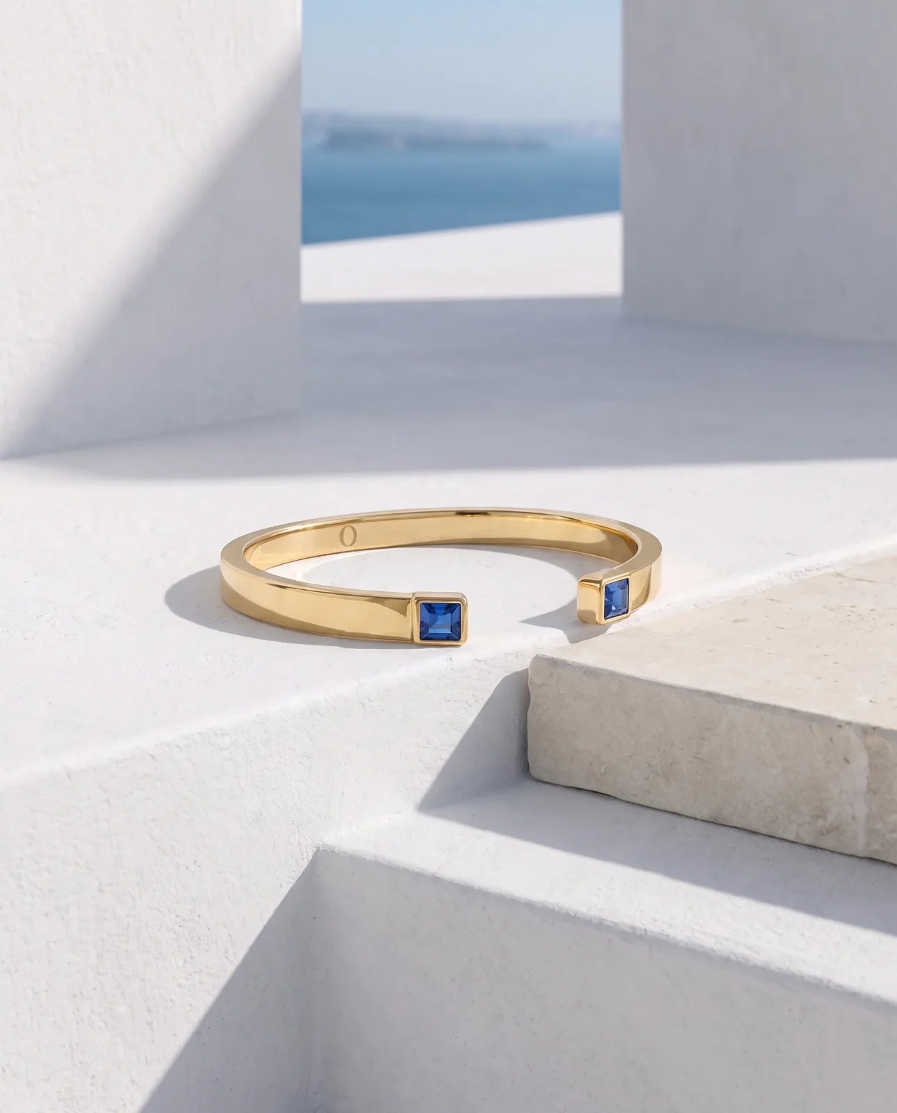 Portico Cuff in 14k yellow gold, a flat polished open cuff with a square step-cut blue sapphire set into each terminal, photographed on sunlit whitewashed steps with a sea view framed by a Santorini opening