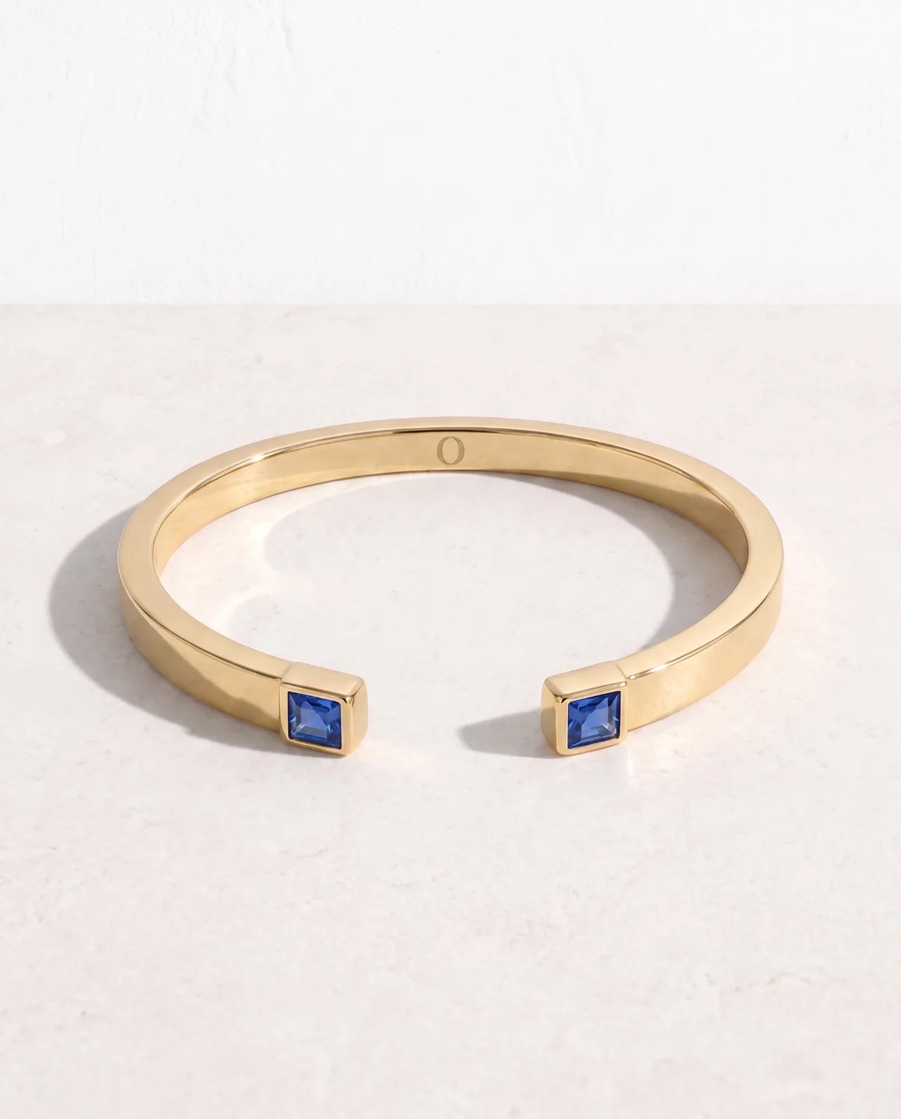 Portico Cuff in 14k yellow gold, a flat polished open cuff with a square step-cut blue sapphire set into each terminal, angled studio shot on a pale stone surface showing both terminals and the inner O engraving