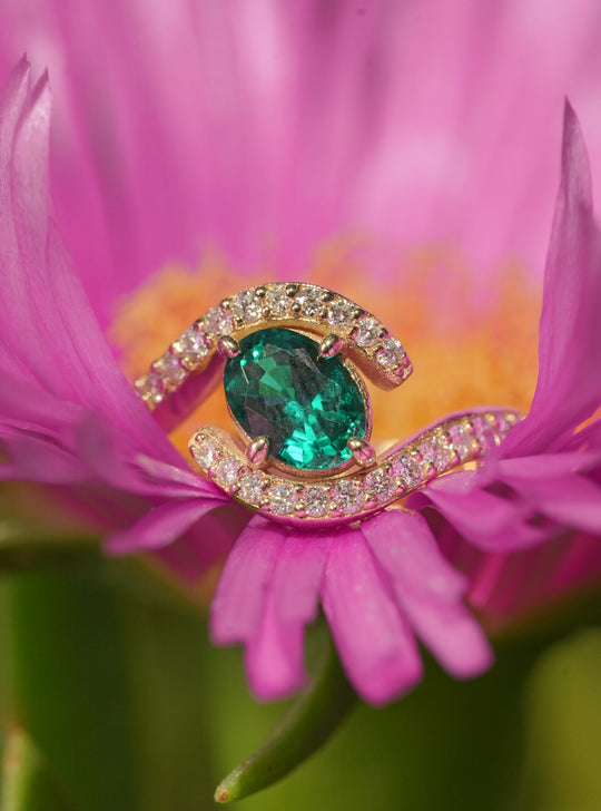 Gold ring with a vibrant emerald center stone and a spiral pavé diamond design, placed on a green leaf background.