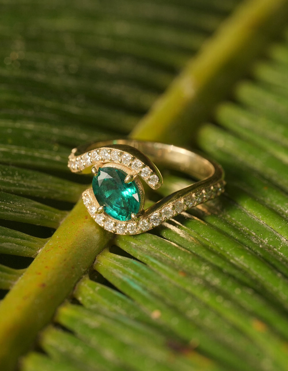 Gold ring with a vibrant emerald center stone and a spiral pavé diamond design, placed on a green leaf background.