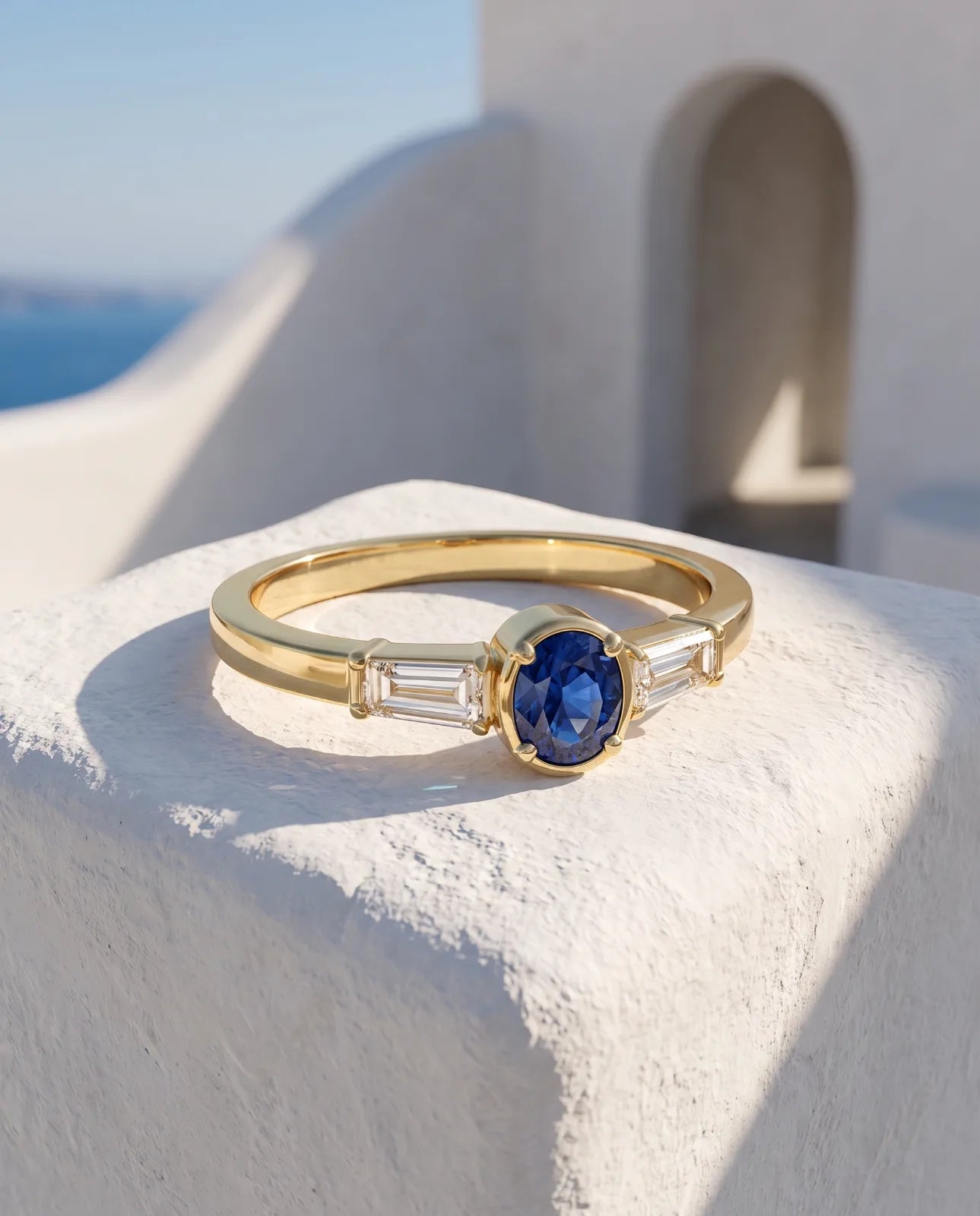 Tholos Ring in 14k yellow gold, an oval blue sapphire set east-west with a baguette diamond on each shoulder, resting on a sunlit whitewashed Santorini ledge with the Aegean sea and a plaster arch behind