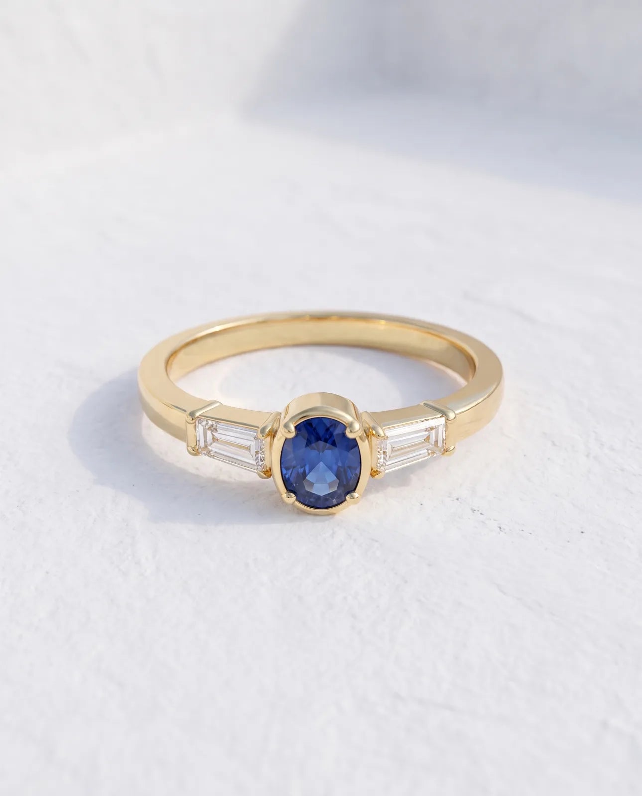 Tholos Ring in 14k yellow gold, an oval blue sapphire set east-west with a baguette diamond on each shoulder, standing upright on a clean white studio surface with soft shadow