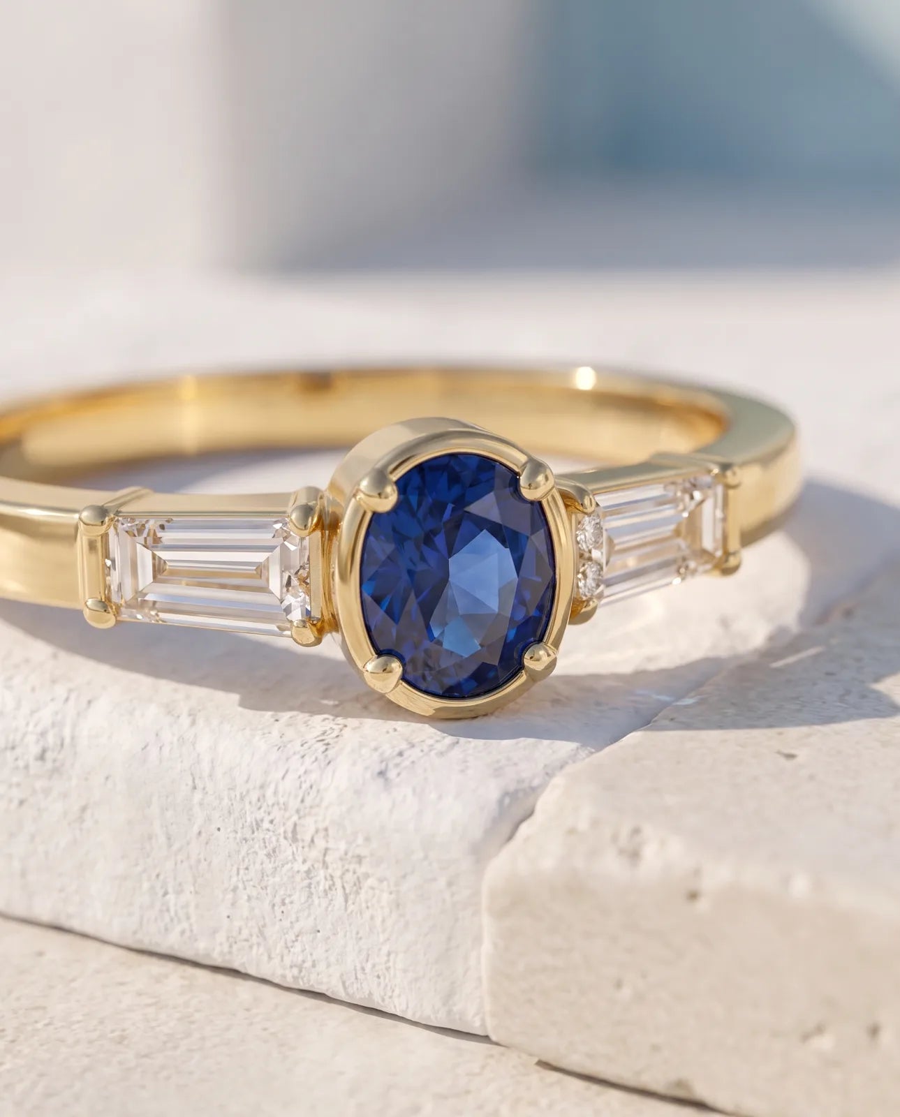 Tholos Ring in 14k yellow gold, an oval blue sapphire set east-west with a baguette diamond on each shoulder, macro detail of the four-prong cradle and bar-set baguettes on a white stone edge