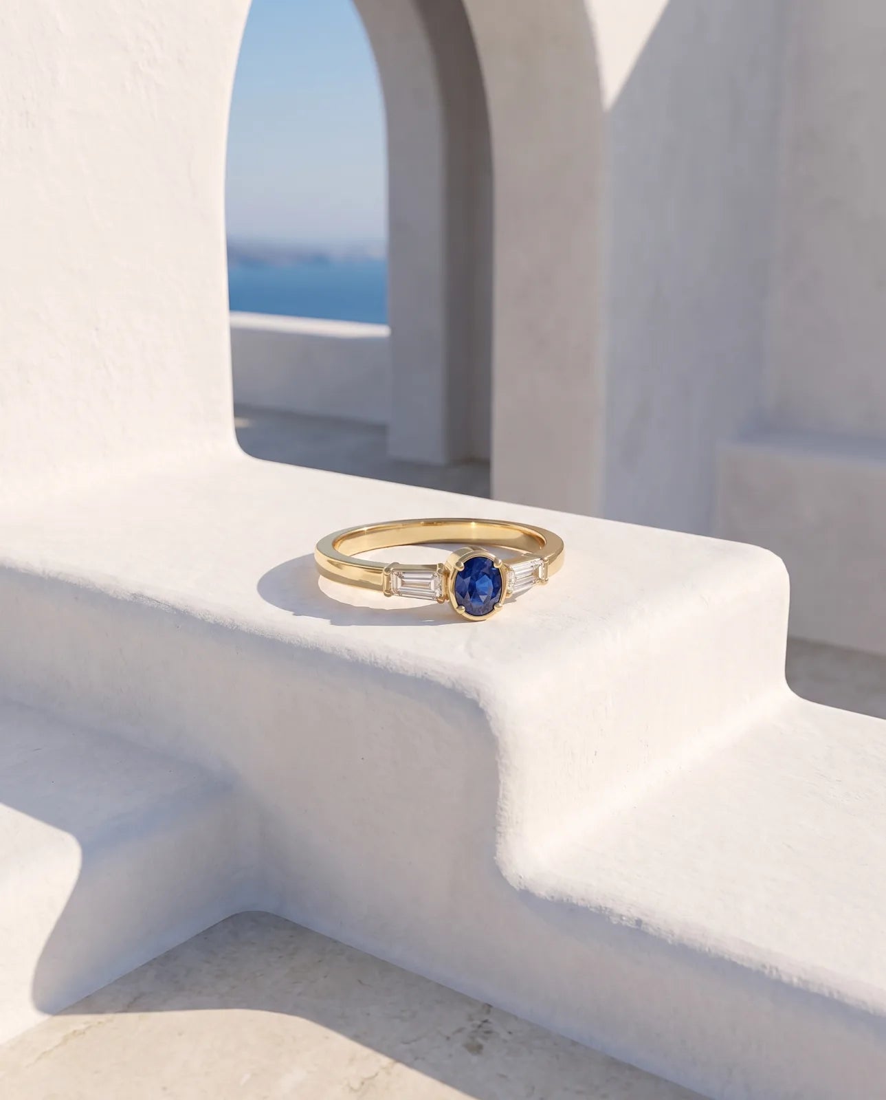 Tholos Ring in 14k yellow gold, an oval blue sapphire set east-west with a baguette diamond on each shoulder, shown small in scale on white Santorini steps beneath a whitewashed arch overlooking the sea