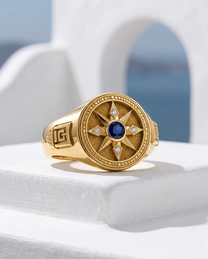 Vesper Signet in 14k yellow gold, a blue sapphire at the centre of an eight-point compass star set with four diamonds, with Greek key shoulders, angled on a whitewashed Santorini ledge with a sunlit arch and the blue caldera behind