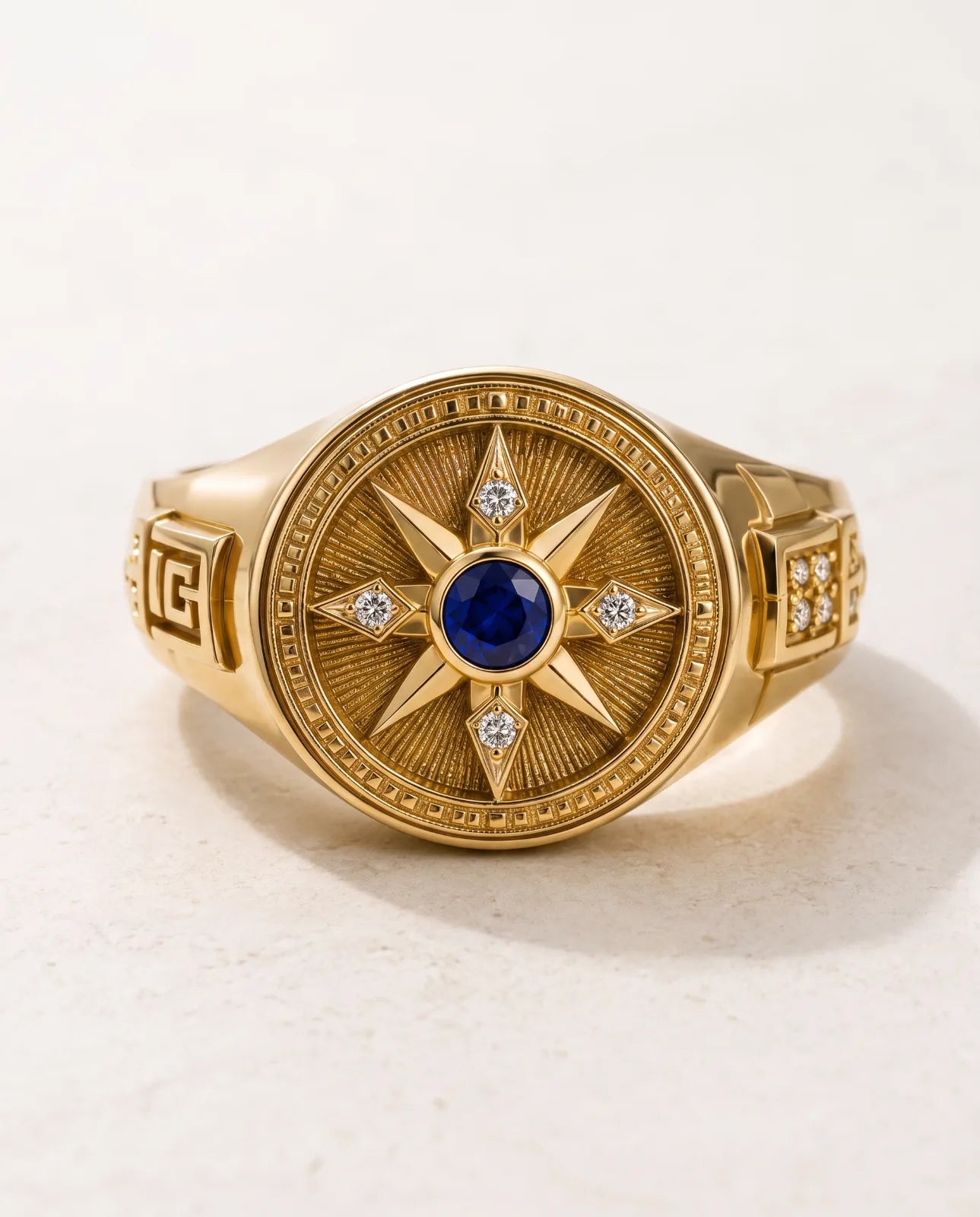 Vesper Signet in 14k yellow gold, a blue sapphire at the centre of an eight-point compass star set with four diamonds, with Greek key shoulders, shown front-on in a clean studio shot on a pale ivory stone background