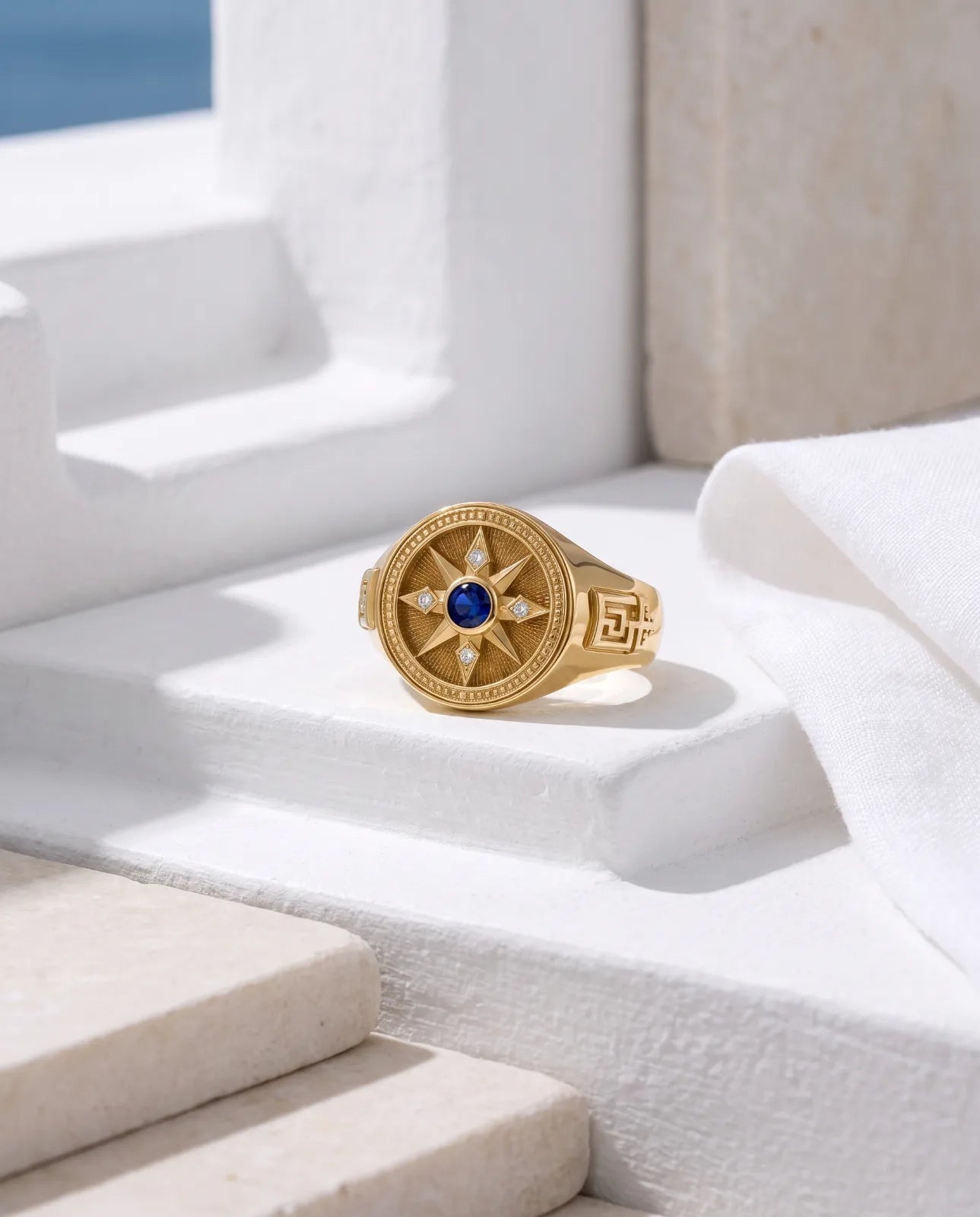 Vesper Signet in 14k yellow gold, a blue sapphire at the centre of an eight-point compass star set with four diamonds, with Greek key shoulders, resting on sunlit whitewashed steps with white linen and the Aegean beyond
