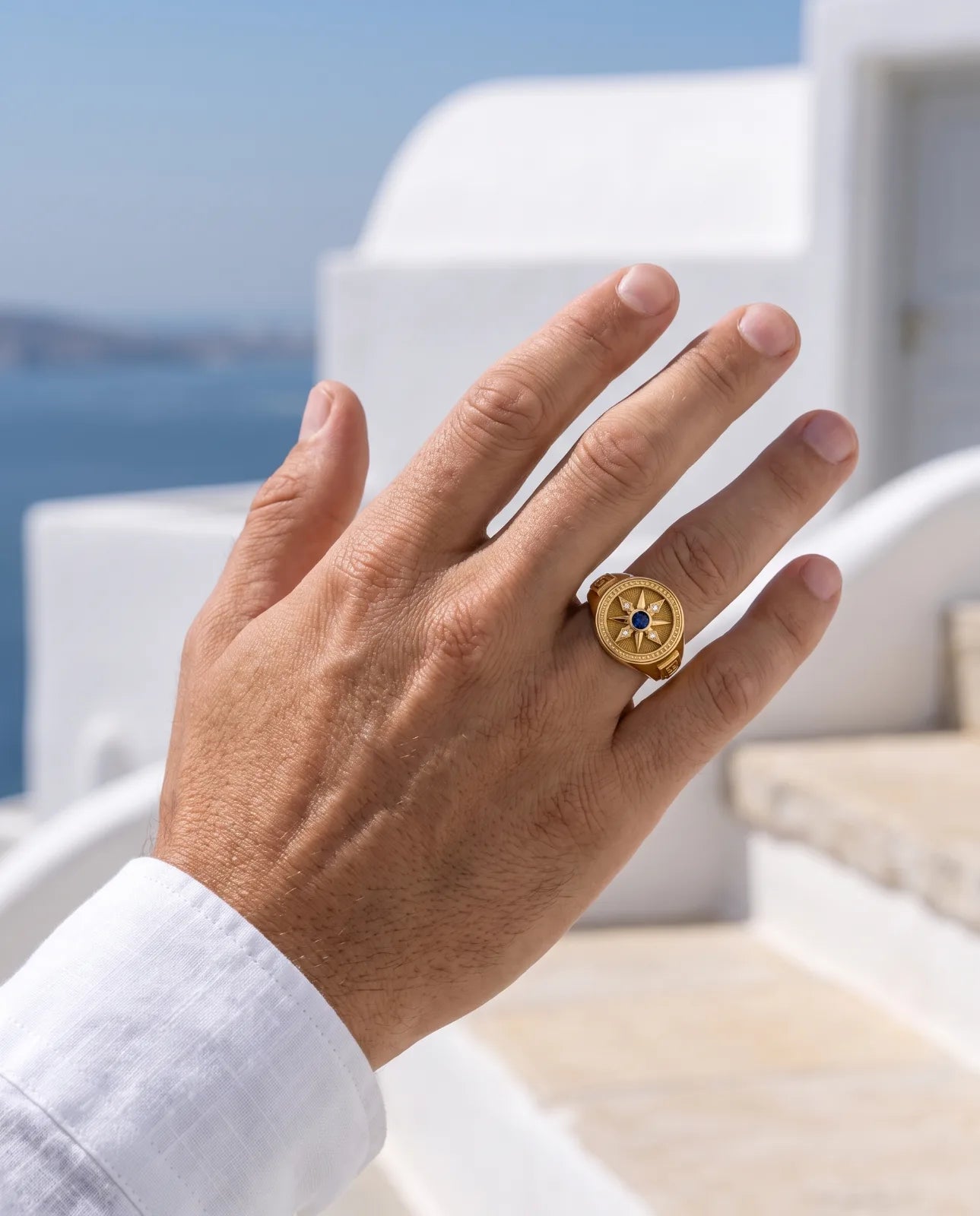 Vesper Signet in 14k yellow gold, a blue sapphire at the centre of an eight-point compass star set with four diamonds, with Greek key shoulders, worn on the middle finger of a hand against whitewashed Santorini walls and the sea